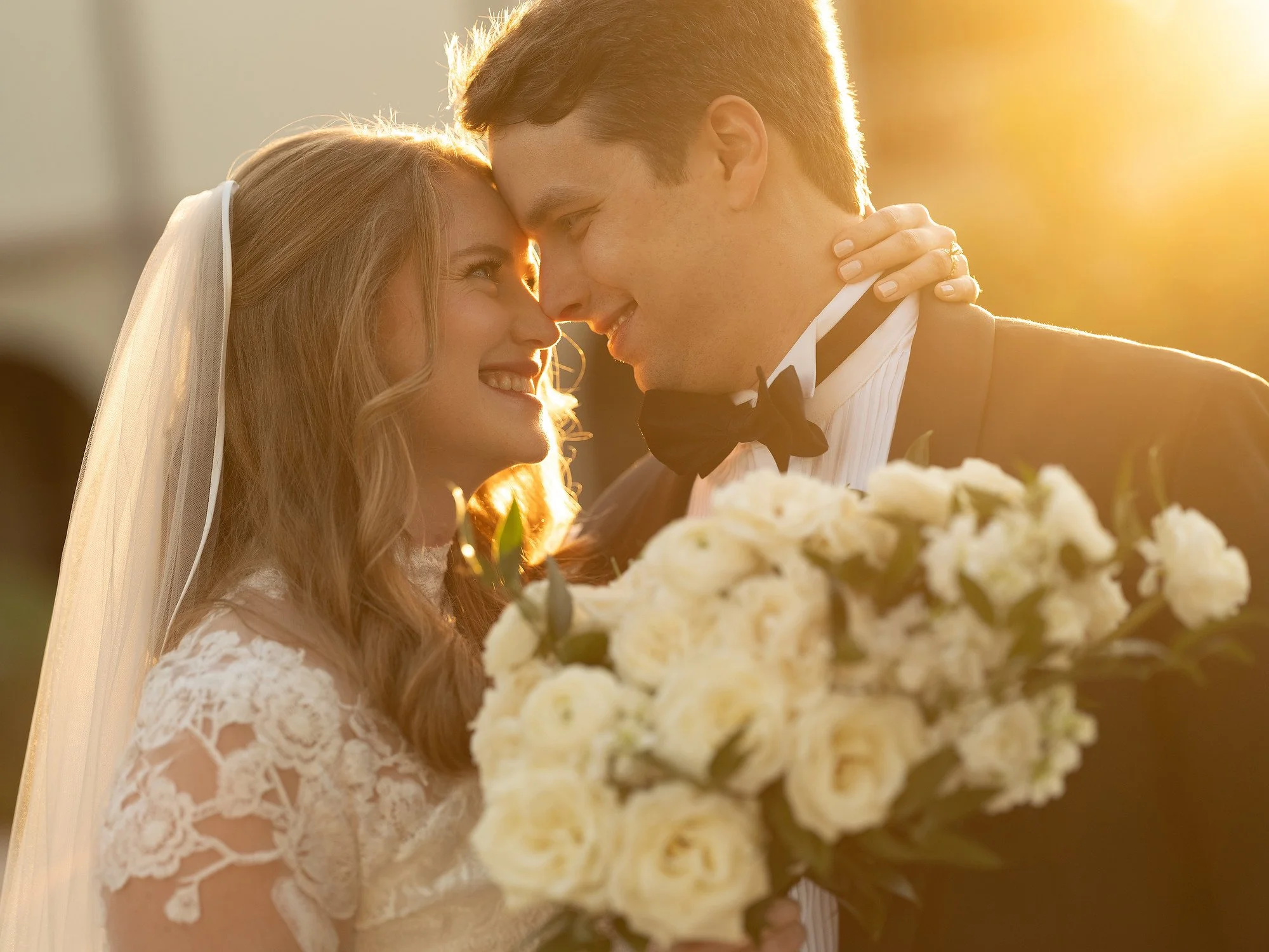 A bride and groom smiling with foreheads touching, holding a wedding bouquet, during sunset.