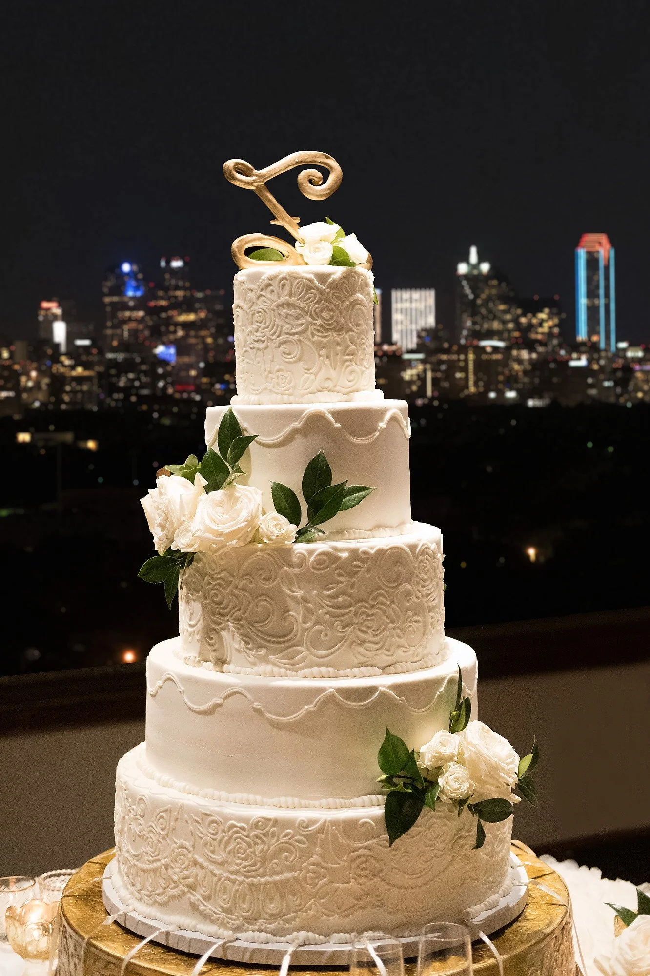 A tall, elegant, five-tier white wedding cake with intricate piping and floral decorations, topped with a golden swirl design and set against a nighttime city skyline.