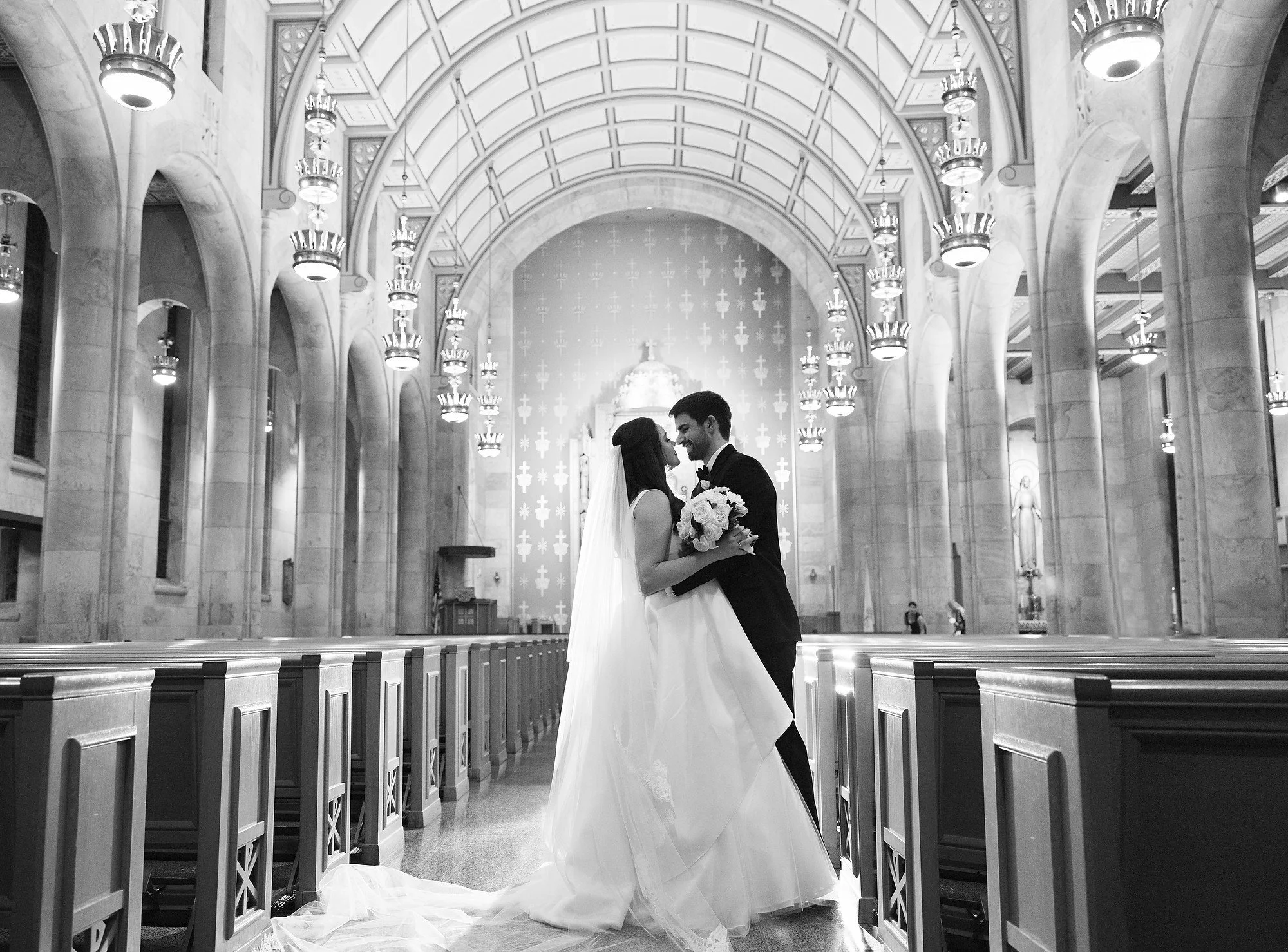 A bride and groom standing close together inside a church, smiling and embracing, with the bride holding a bouquet of flowers.