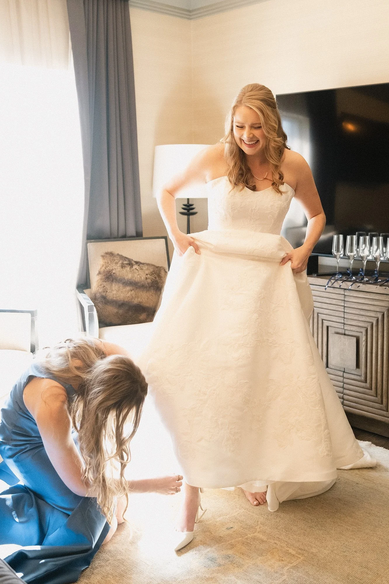 A bride in a wedding dress is smiling while trying on shoes with the help of a woman in a blue dress, in a well-lit room with a patterned carpet, a sofa, and a side table with champagne flutes.
