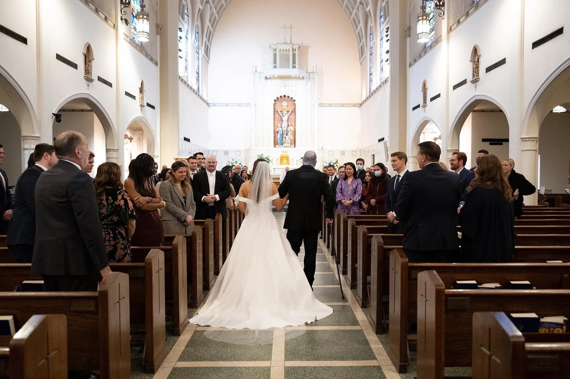 A bride and an elderly man, likely her father, walking down the aisle of a church during a wedding ceremony, with guests on either side watching.
