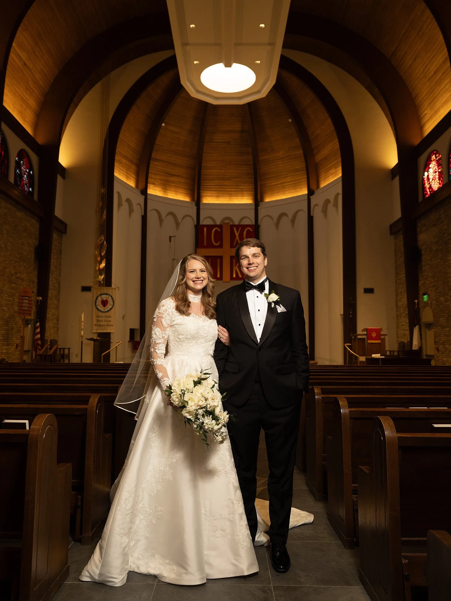 A bride and groom standing together inside a church, smiling at the camera, with pews and religious symbols in the background.