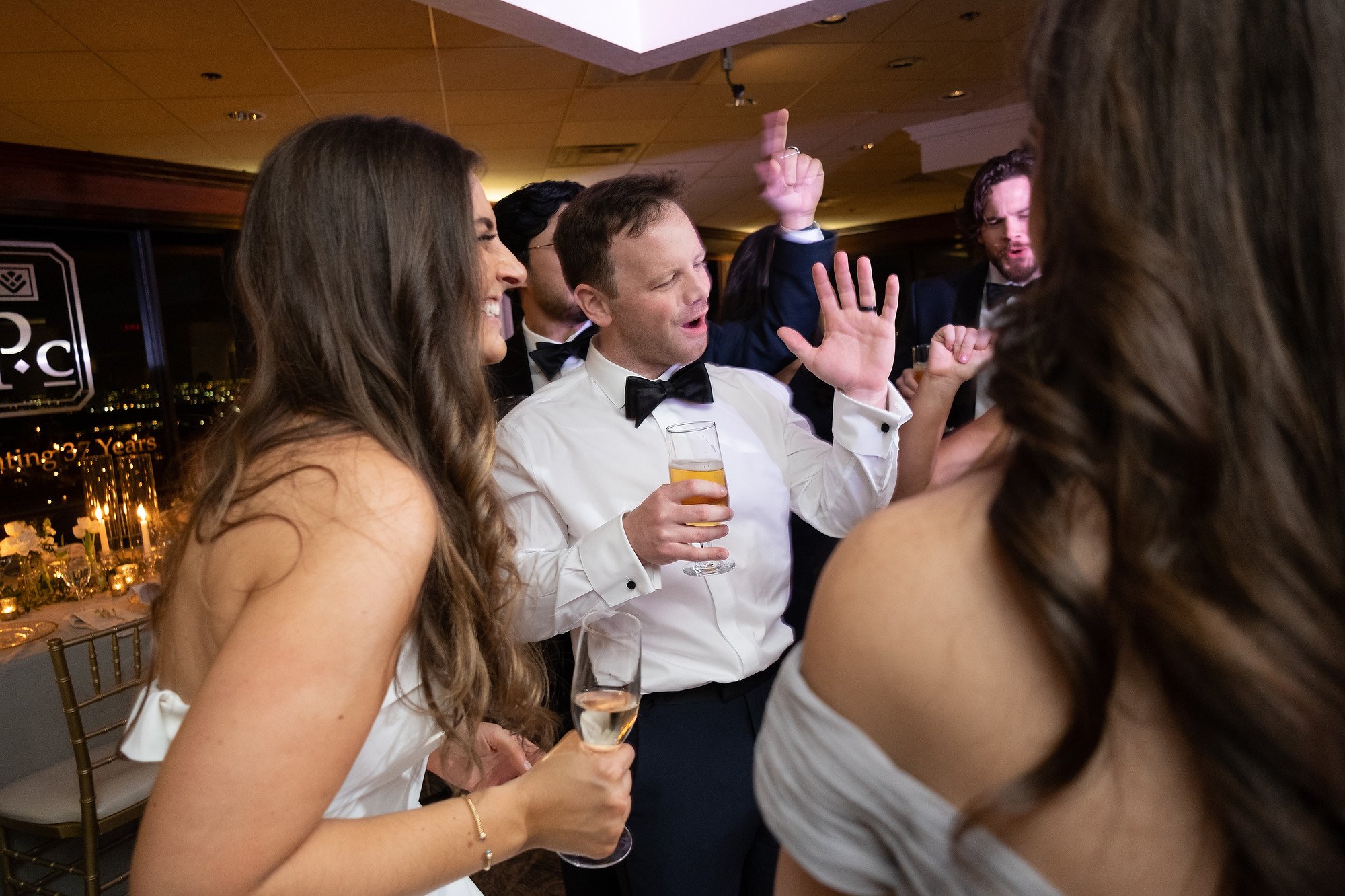 Group of people dancing and celebrating at a formal event, with drinks in hand, in an indoor banquet hall.