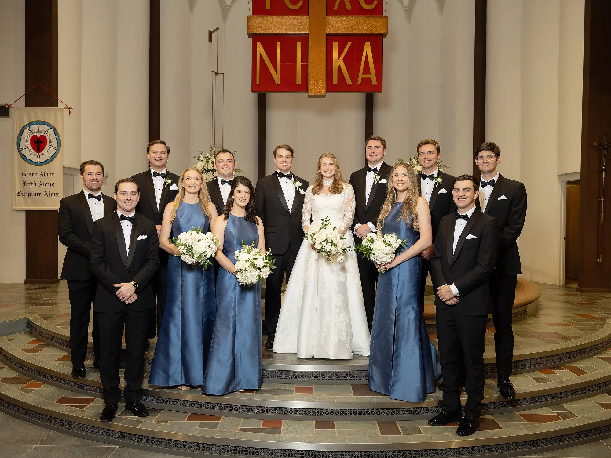 Group wedding photo with bride and groom in center, surrounded by bridesmaids and groomsmen in church.