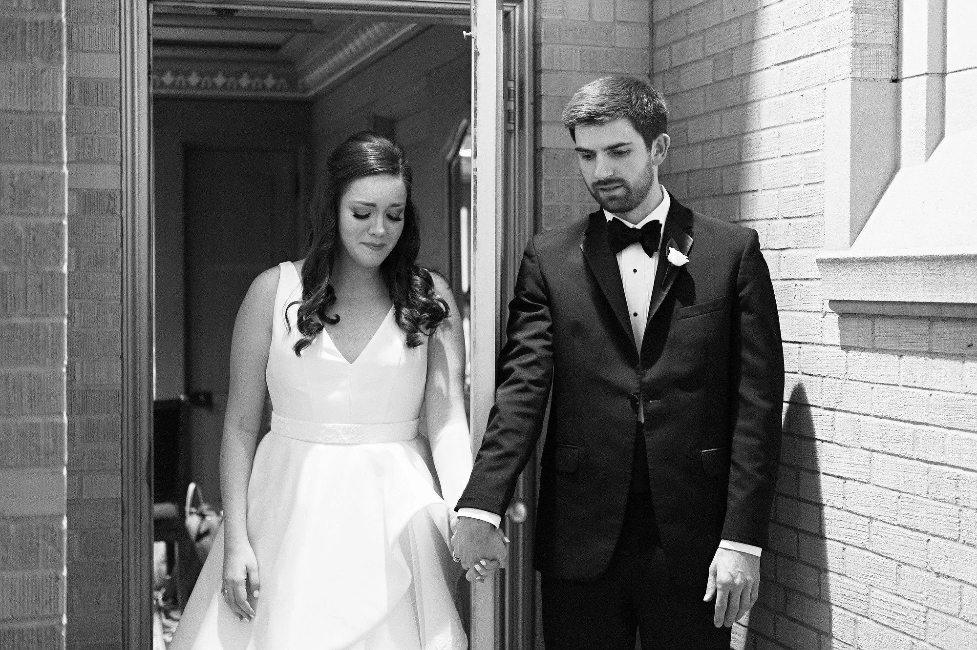 Black and white photo of a bride and groom holding hands with eyes closed, standing outdoors near a brick wall.