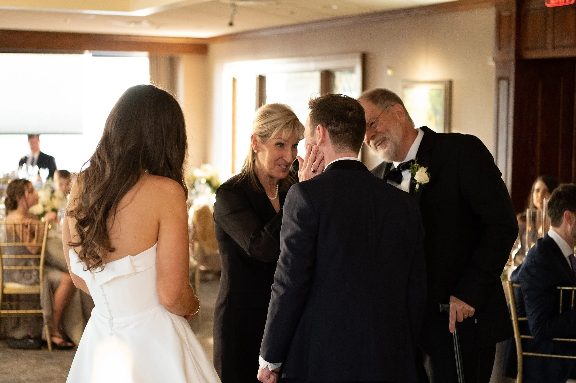 A wedding reception scene where a woman is whispering into a man's ear while an older man smiling adjacent, and a woman in a white dress stands nearby in a decorated banquet hall.