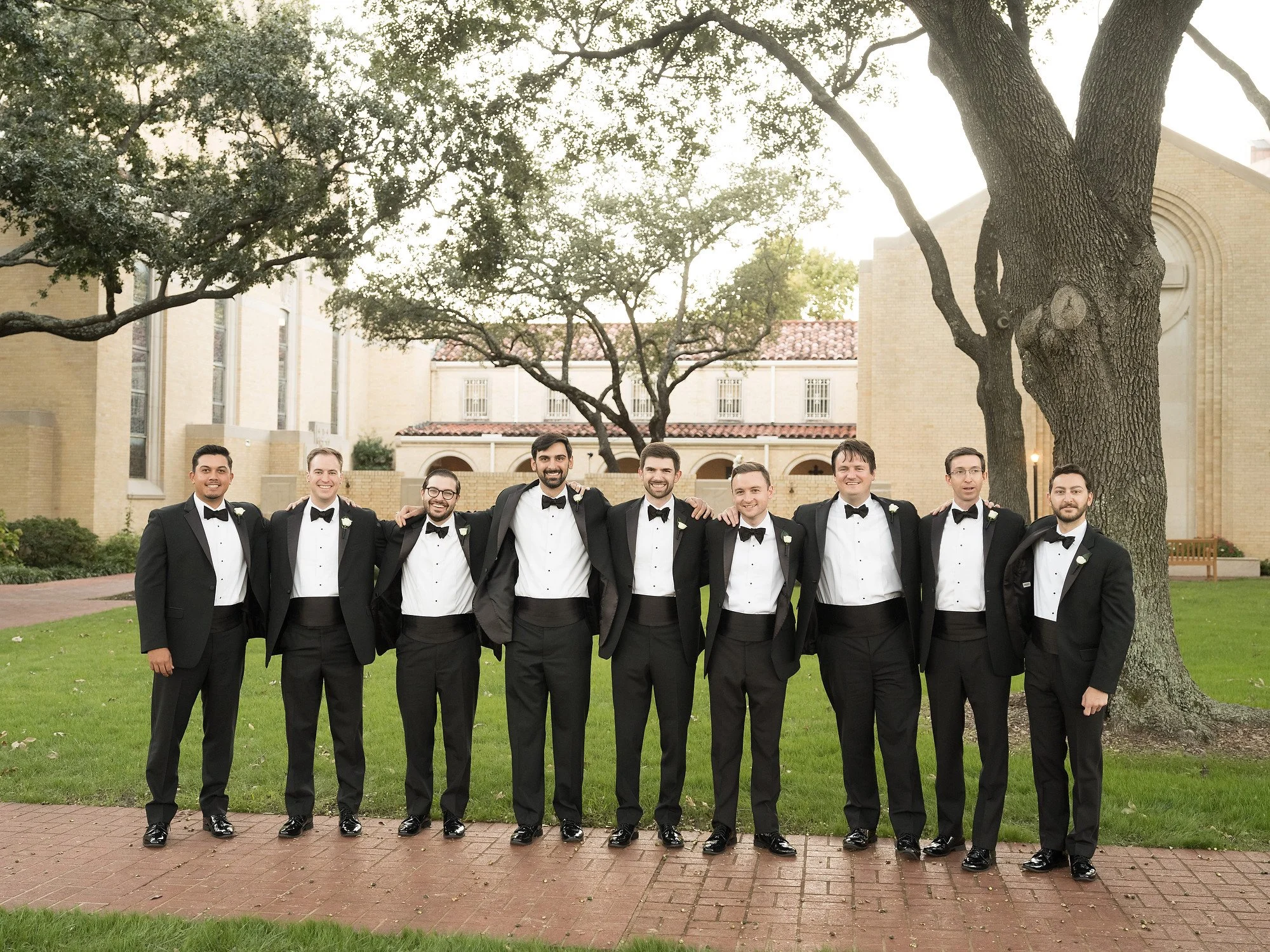 A group of nine men in black tuxedos and bow ties standing outdoors in front of trees and a building, smiling with arms around each other's shoulders.