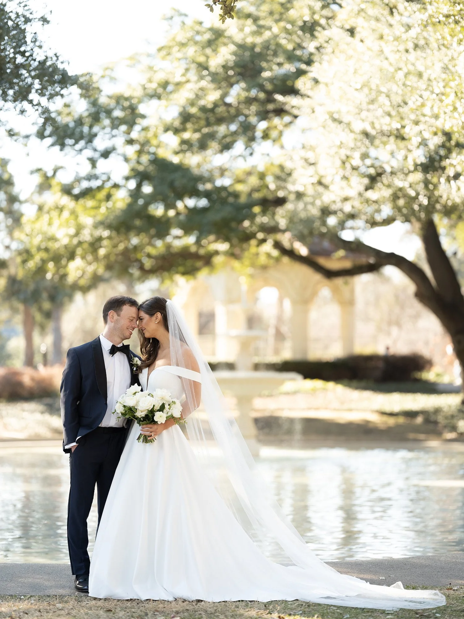 A bride and groom standing close together outdoors, smiling and touching foreheads, with the bride holding a bouquet of white flowers, surrounded by trees and a fountain in the background.