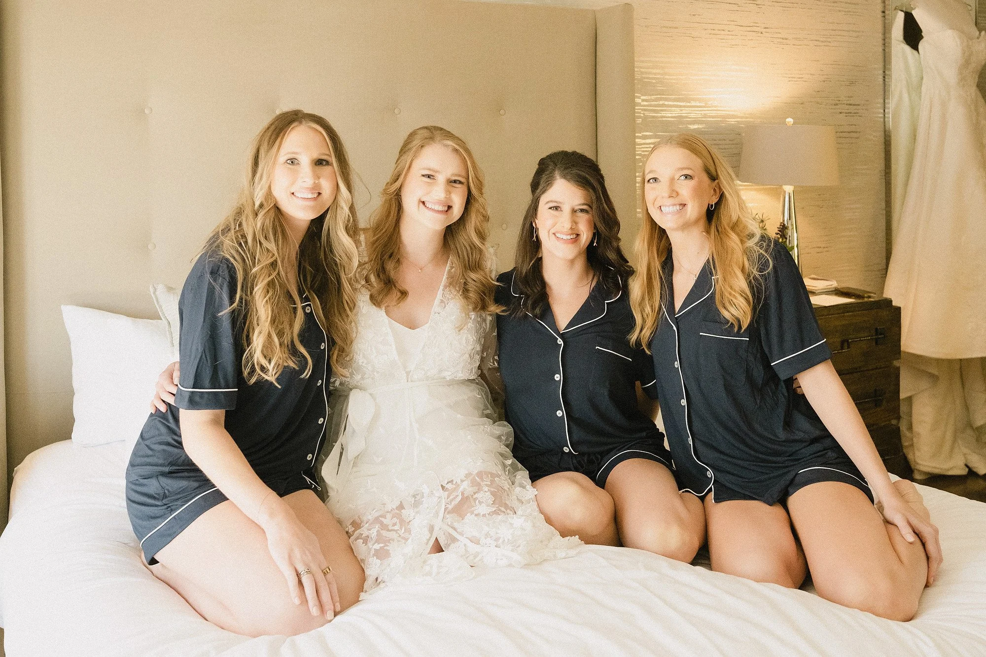 Bride and bridesmaids sitting on a bed, smiling, in a hotel room with a white bedsheet, beige headboard, and nightstand with lamp.