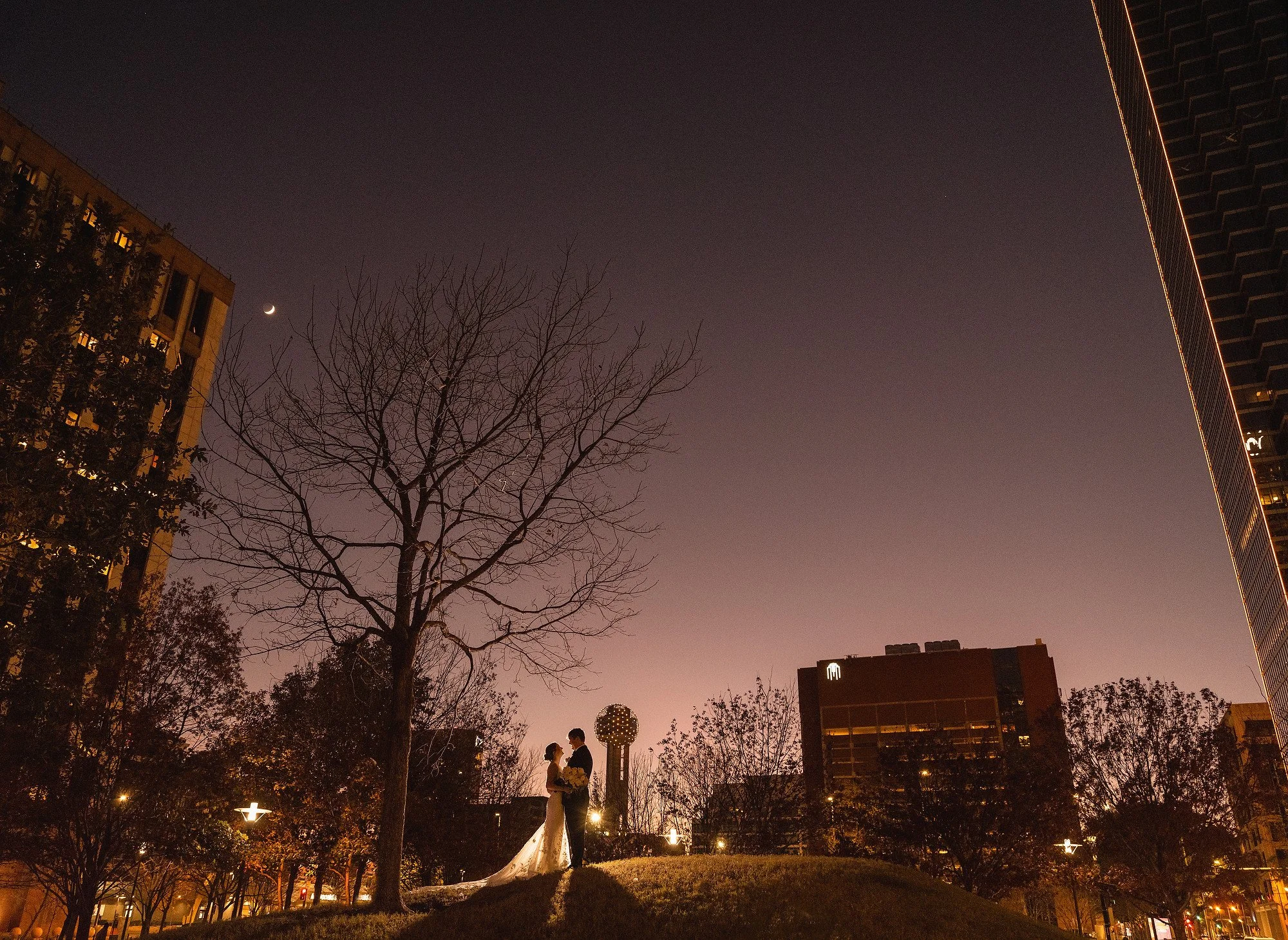 A couple in wedding attire standing on a small hill at dusk, with empty trees and city buildings around them, under a darkening sky with a crescent moon.