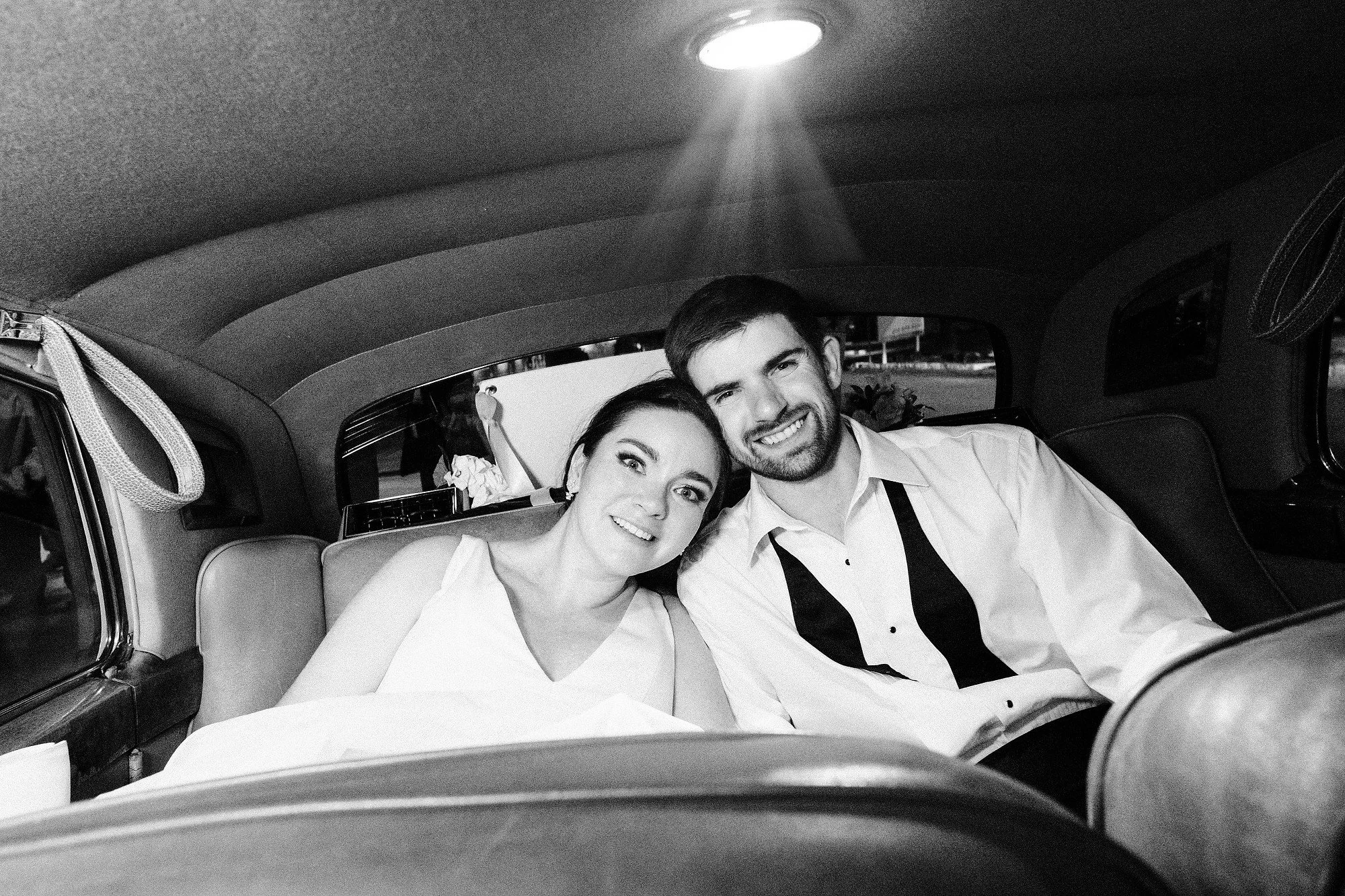 A smiling couple sitting close together in the back seat of a vintage car, illuminated by an interior light, in black and white.