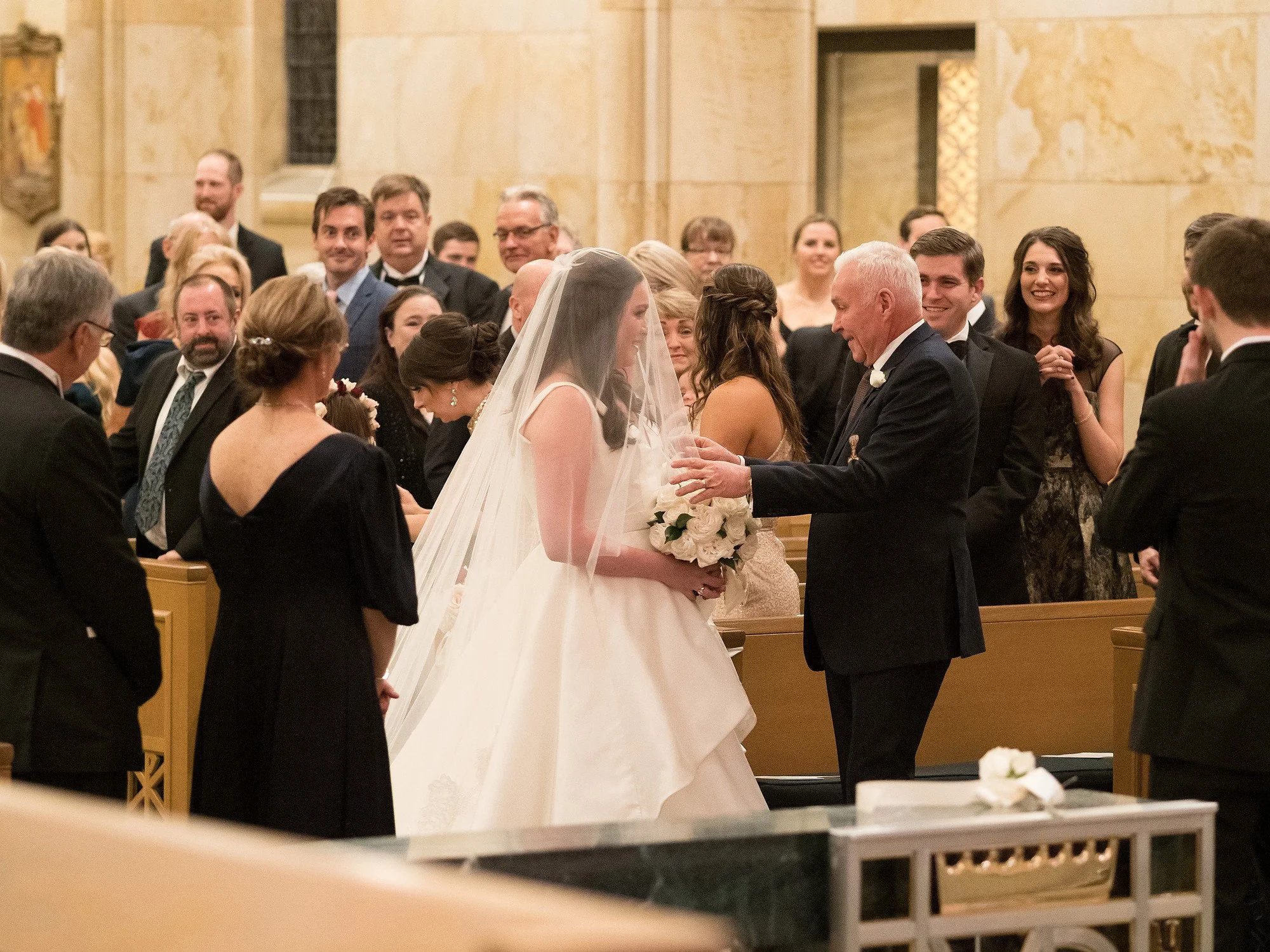 A bride in a wedding dress and veil holding a bouquet of white roses stands next to an older man, possibly her father, who is smiling and extending his hand towards her during a wedding ceremony in a church filled with guests.