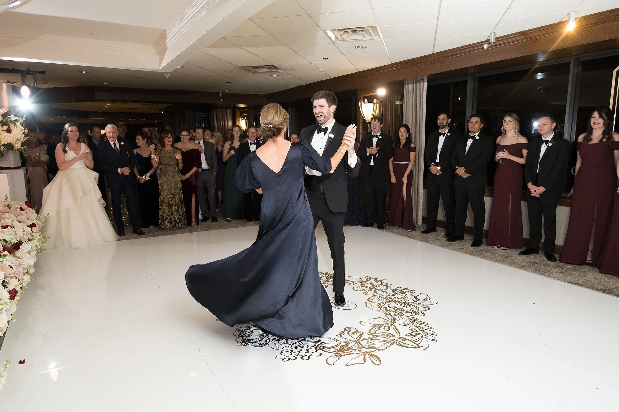 A bride and groom dance in the center of a wedding reception with guests around watching and smiling.
