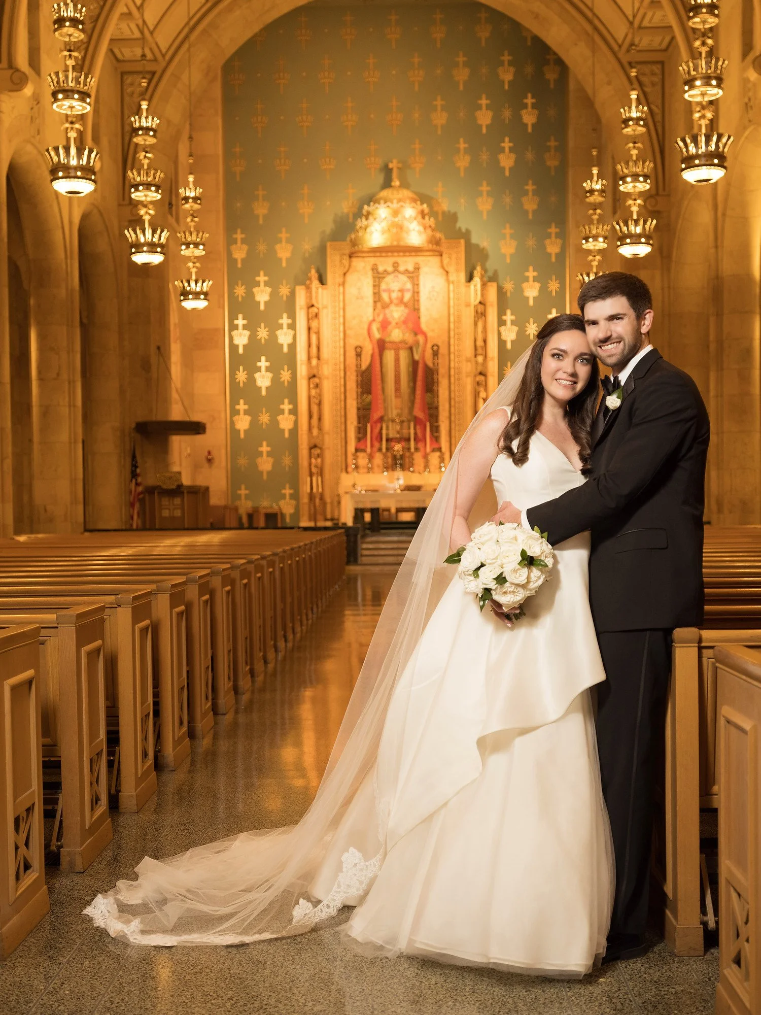 A bride and groom standing together inside a church with ornate decor in the background, smiling at the camera. The bride is in a white wedding gown with a long veil and holding a bouquet of white roses. The groom is in a black tuxedo with a white sh