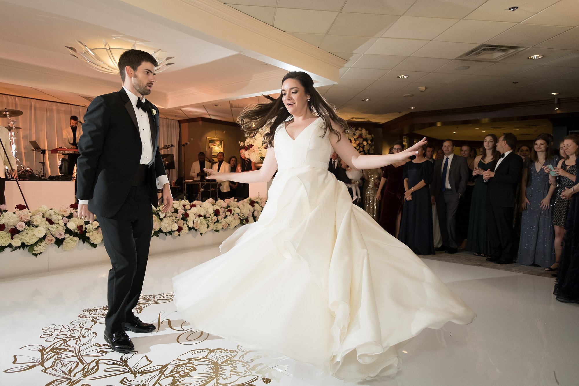 A bride in a flowing white wedding gown spins on the dance floor with her left arm extended, while a groom in a black tuxedo looks on at a wedding reception. Guests and a live band are visible in the background, and the dance floor is decorated with 