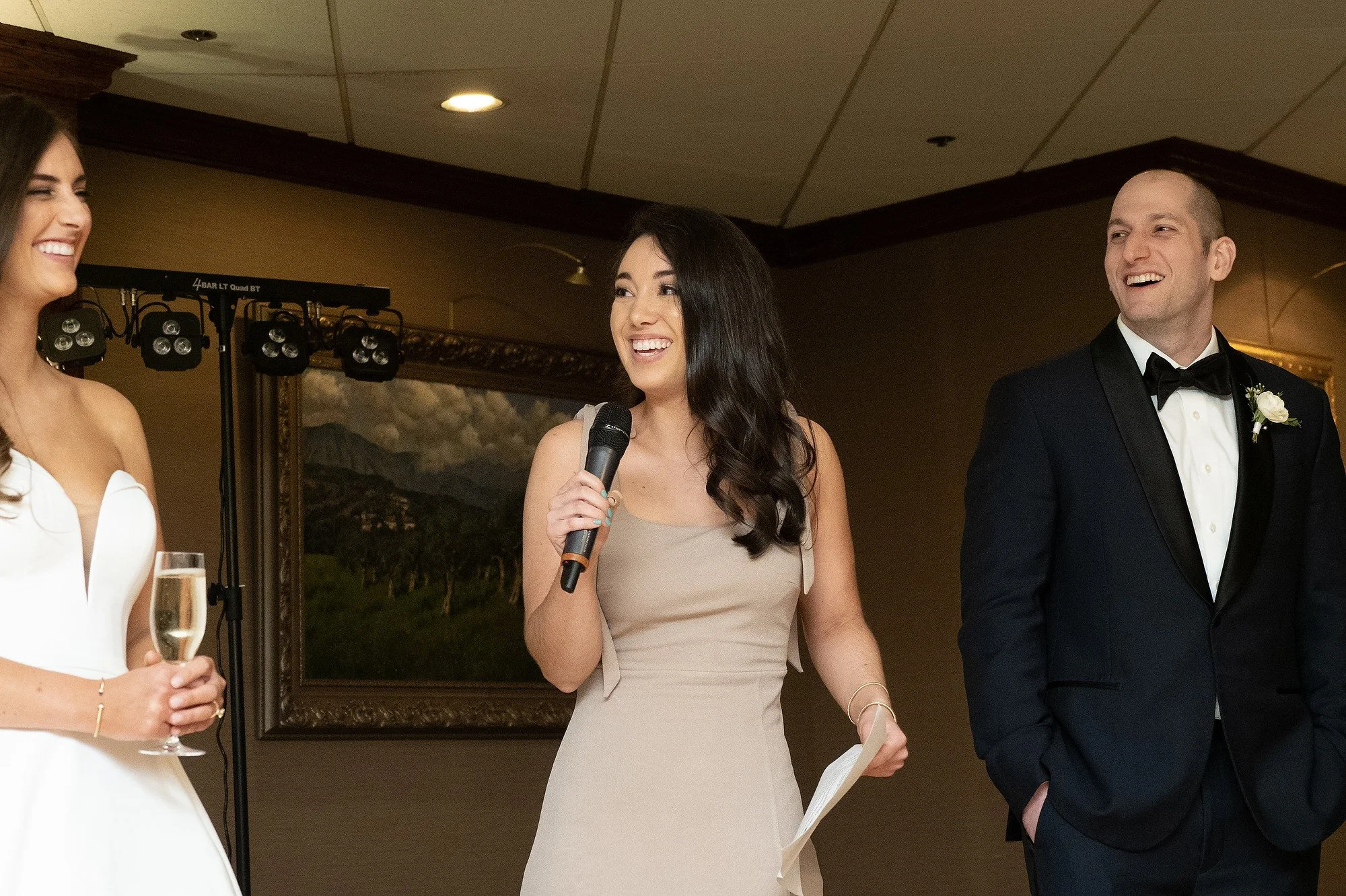 A woman wearing a beige dress holding a microphone and a paper, standing next to a man in a black tuxedo with a boutonniere, at a wedding reception.
