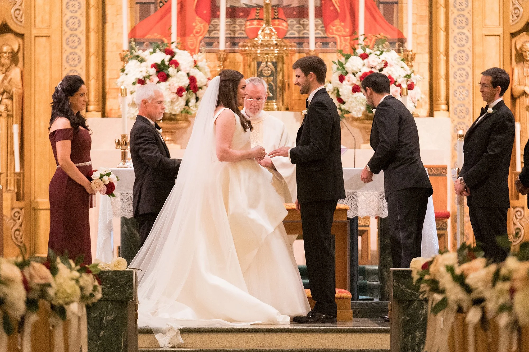 A bride and groom exchange vows at an altar during a wedding ceremony, surrounded by attendants in a church decorated with flowers.
