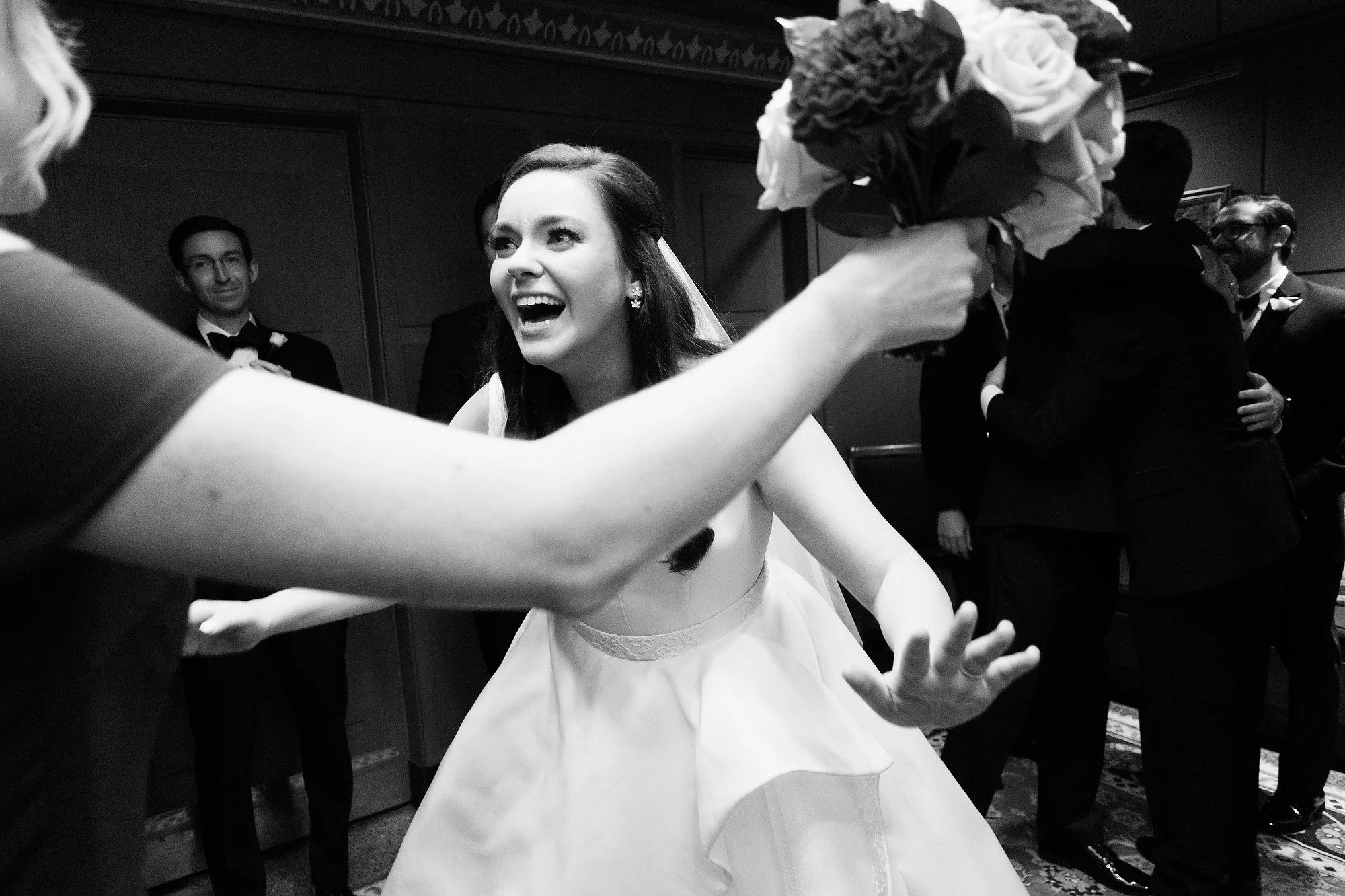 A joyful woman in a wedding dress smiling and reaching out to receive a bouquet of flowers at her wedding reception, with bridesmaids and groomsmen in tuxedos in the background.