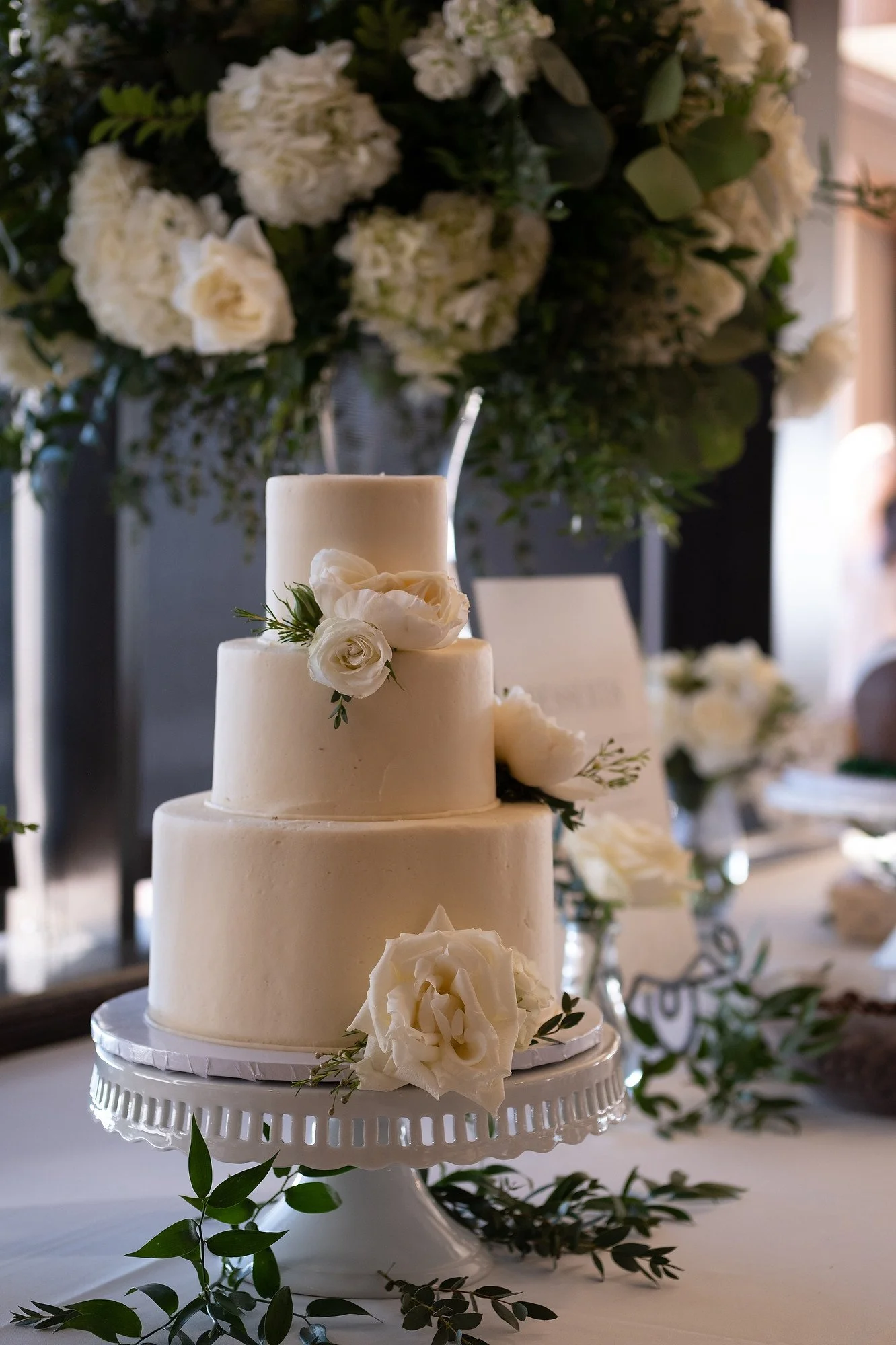 Three-tiered white wedding cake decorated with white roses and greenery, placed on a stand at a wedding reception.