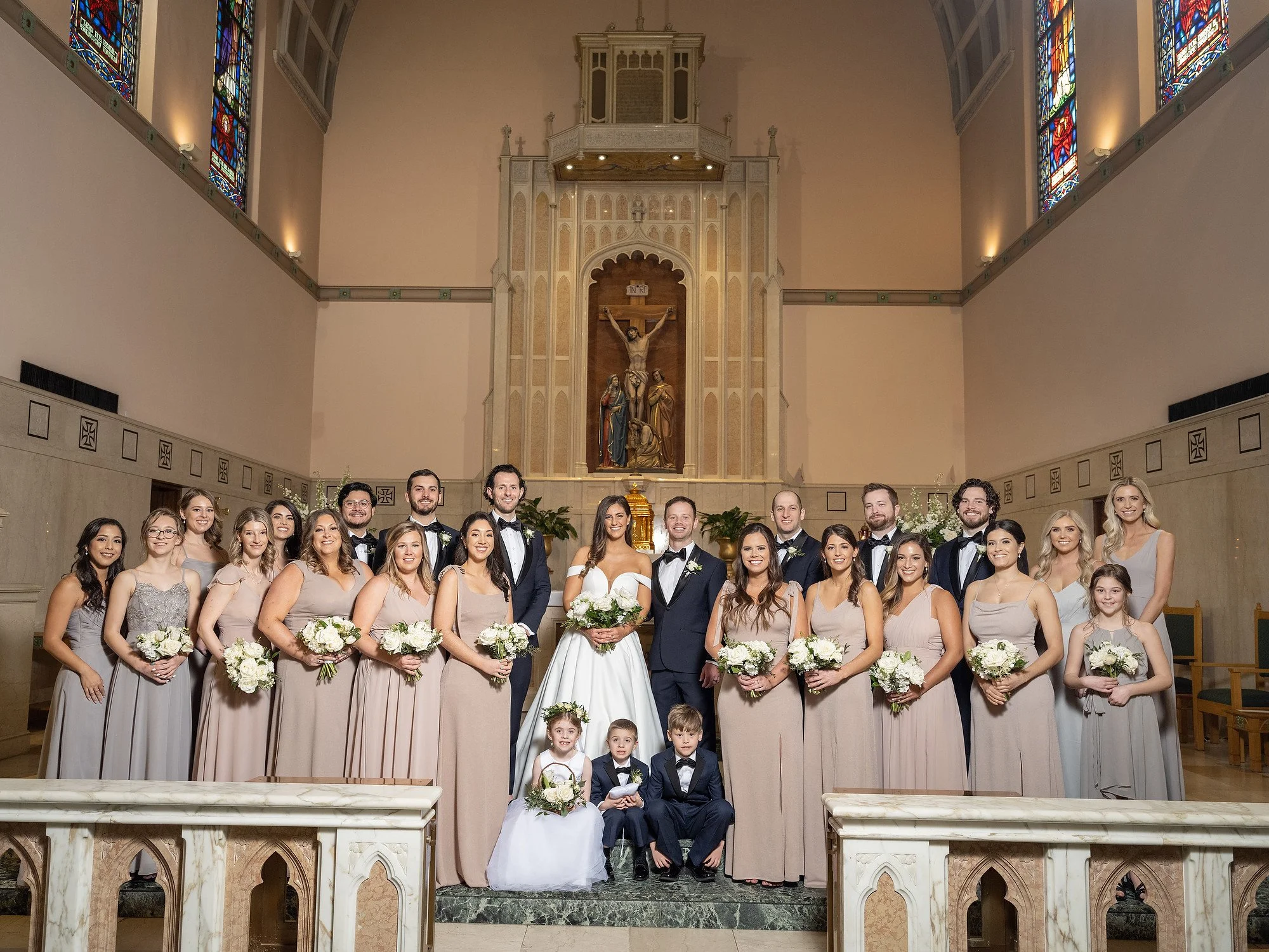 A large wedding party in a church with stained glass windows and religious statues, including the bride and groom at the center, surrounded by bridesmaids, groomsmen, and children, all dressed in formal wedding attire.