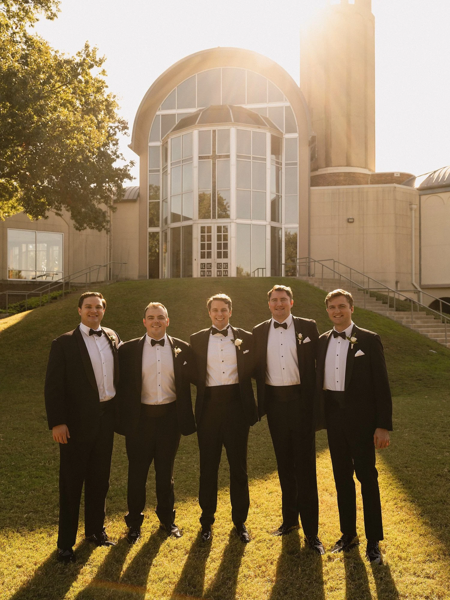 Six men in tuxedos standing outdoors on grass, smiling, with a large glass-fronted building in the background, sunlight shining down.
