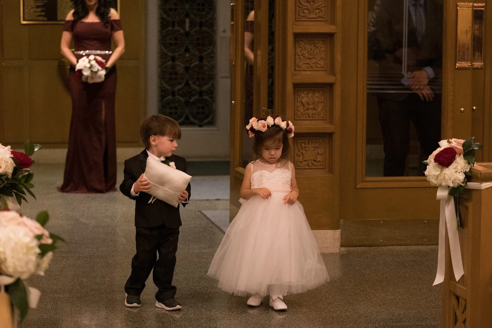 Two young children, a girl in a white dress with a floral headband and a boy in a black tuxedo, are standing in a decorated room, likely at a wedding. The girl looks down while the boy holds a pillow, and a woman in a maroon dress holding a bouquet s