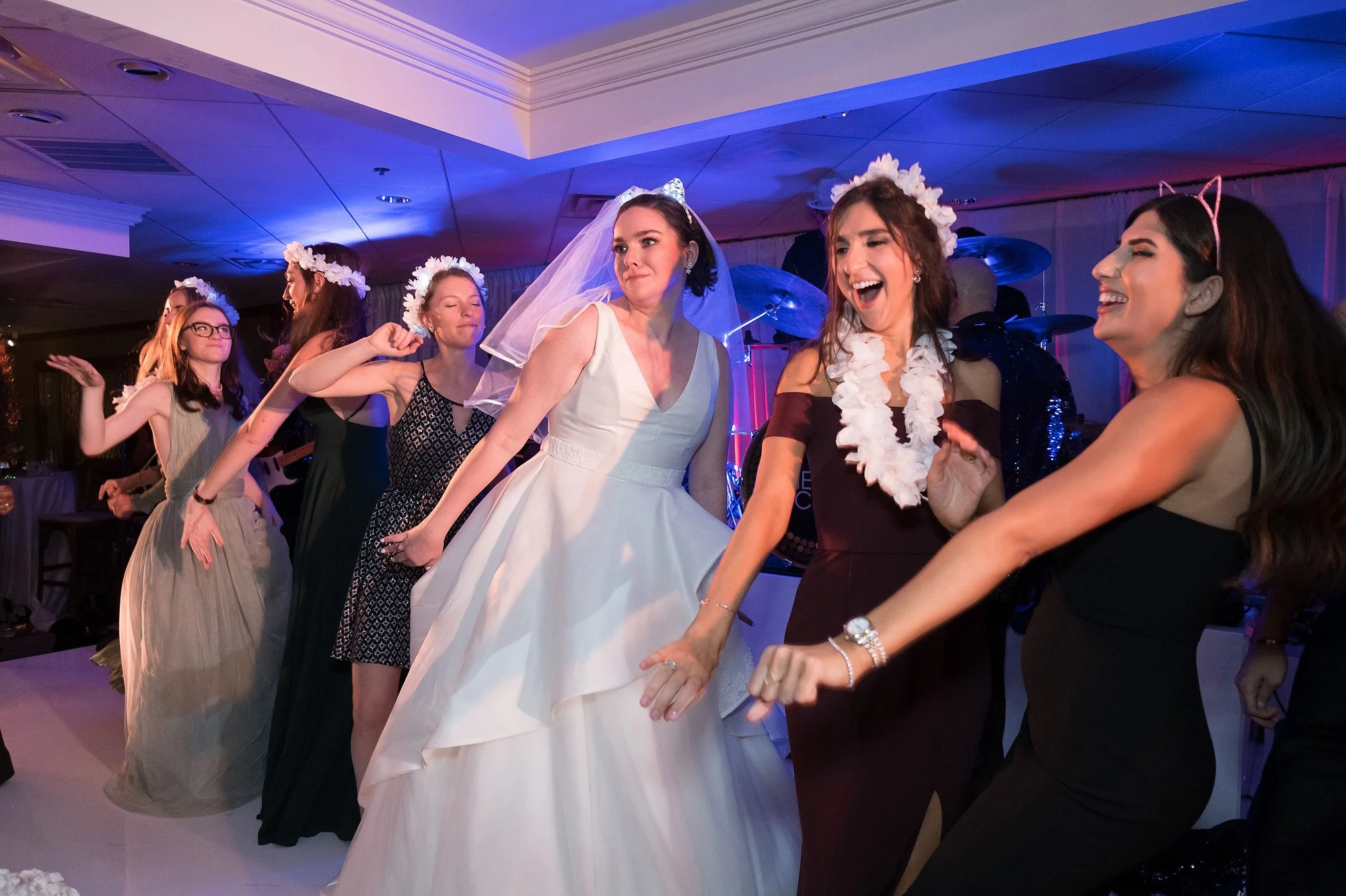 Women dancing at a celebration, with some wearing floral headbands; one woman in a white wedding dress, others dressed in formal attire, some with cat ear headbands. The setting appears to be indoors with colorful lighting.