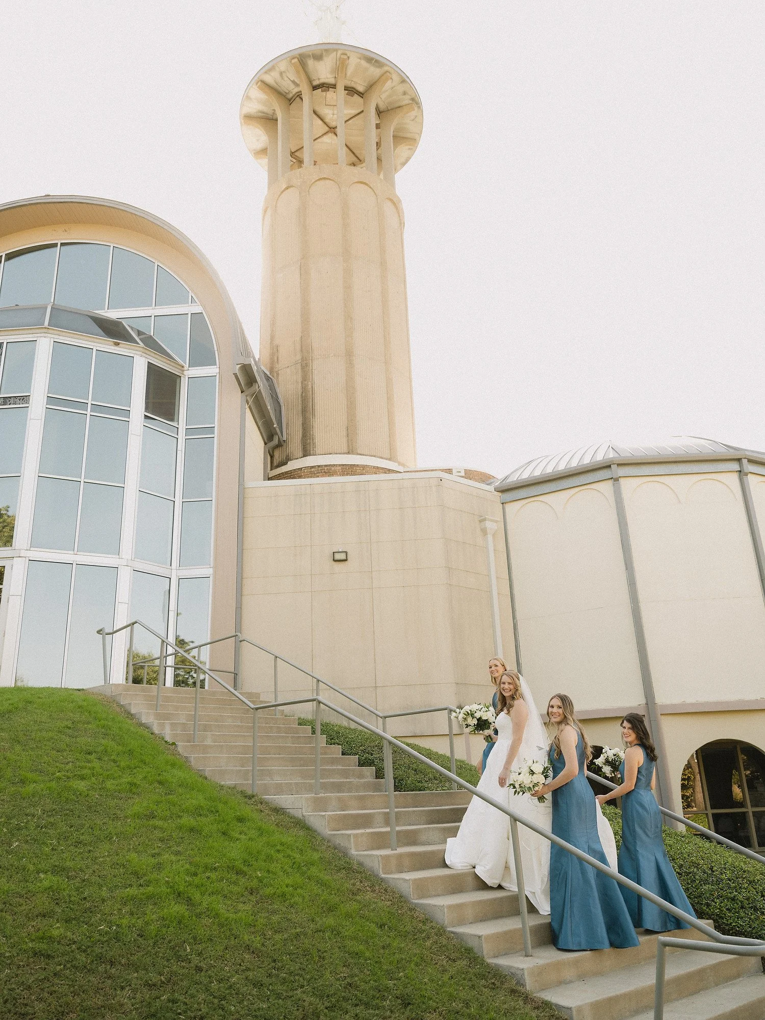 A bride and three bridesmaids walking down outdoor stairs in front of a modern church with glass windows and a tall tower, during daytime.