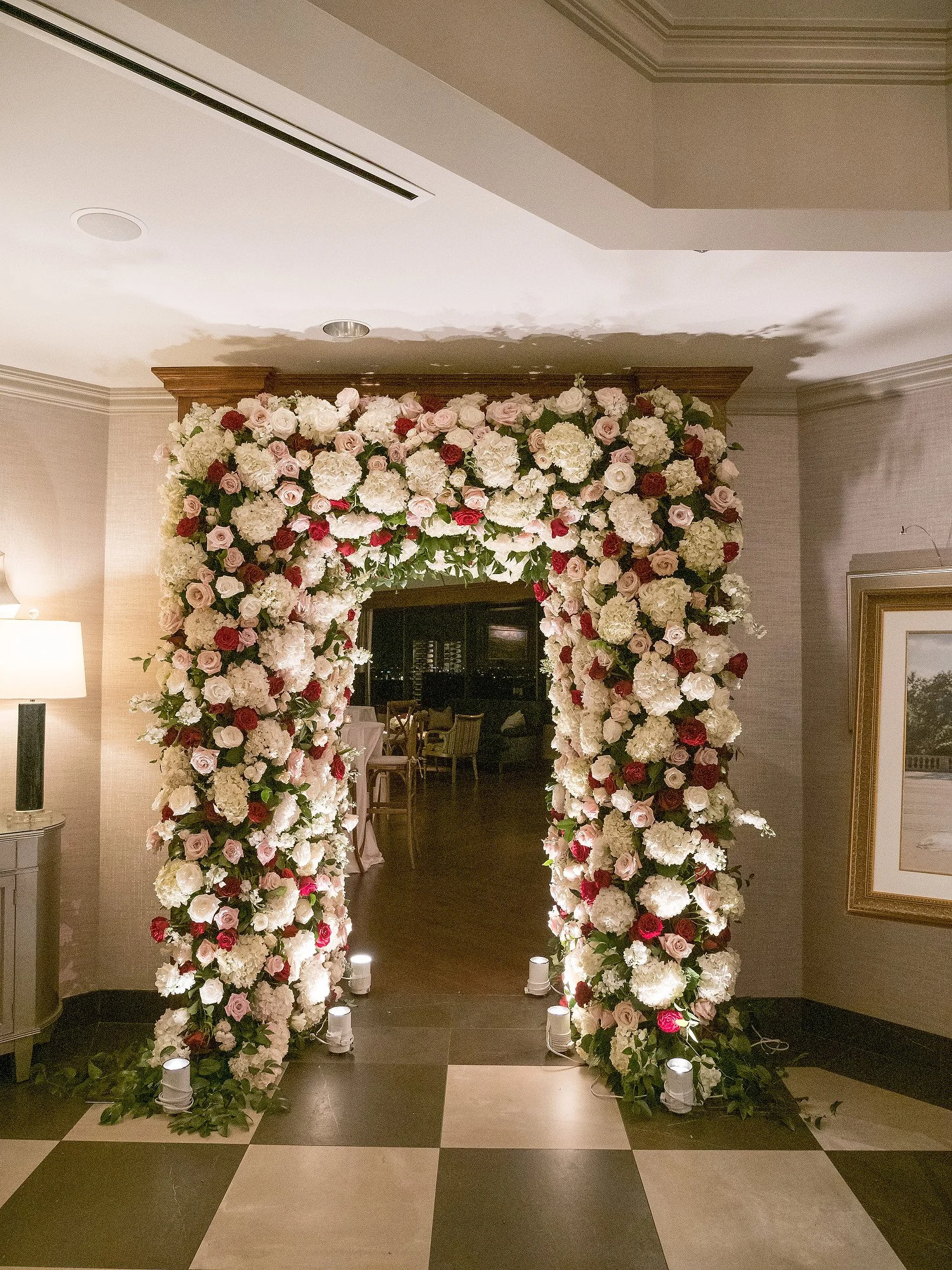 A floral arch made of white, pink, and red roses and hydrangeas set up indoors, illuminated by small ground lights at its base.