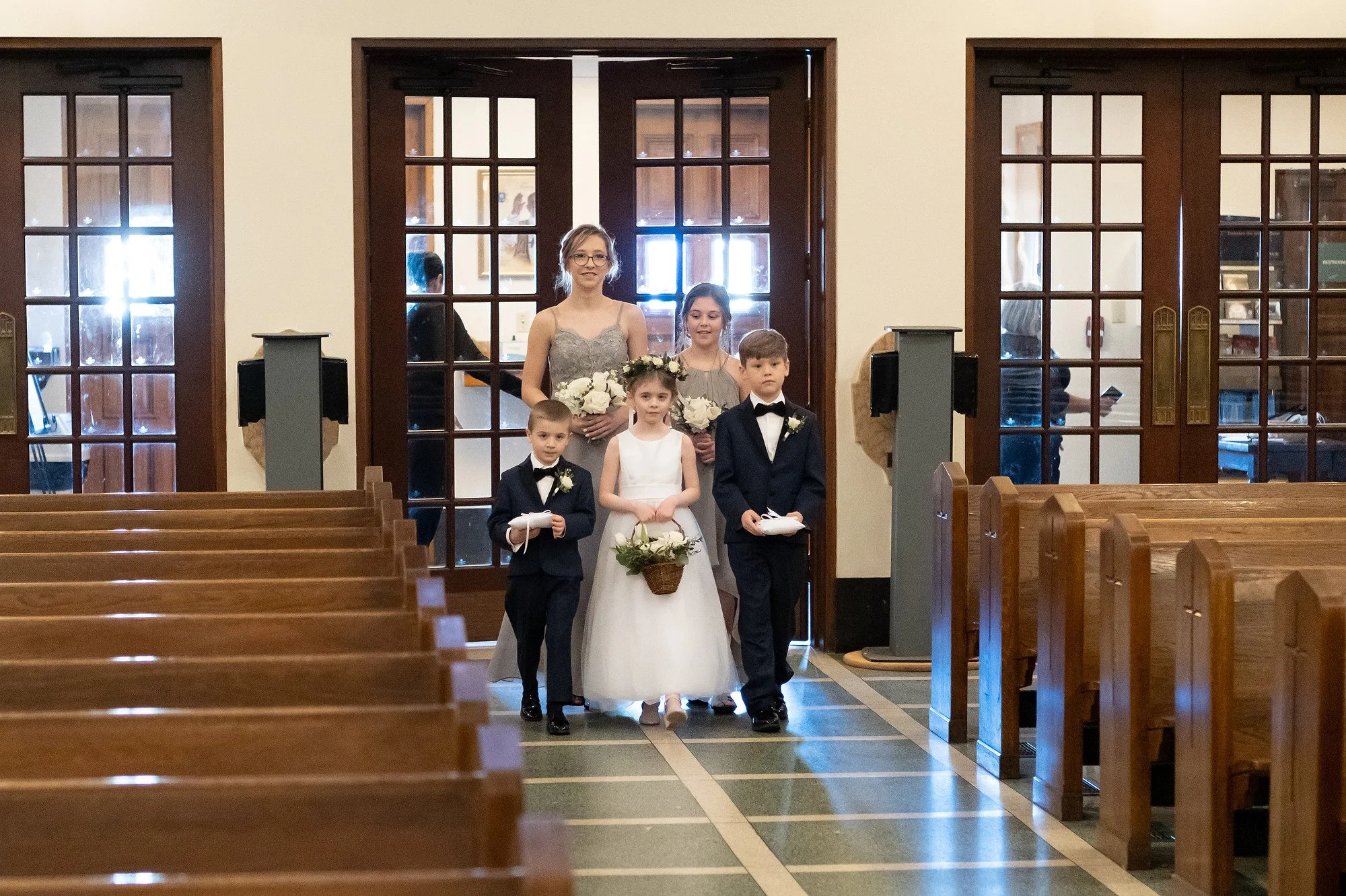 Children and women participating in a wedding procession inside a church, with pews on either side and large glass doors behind them.