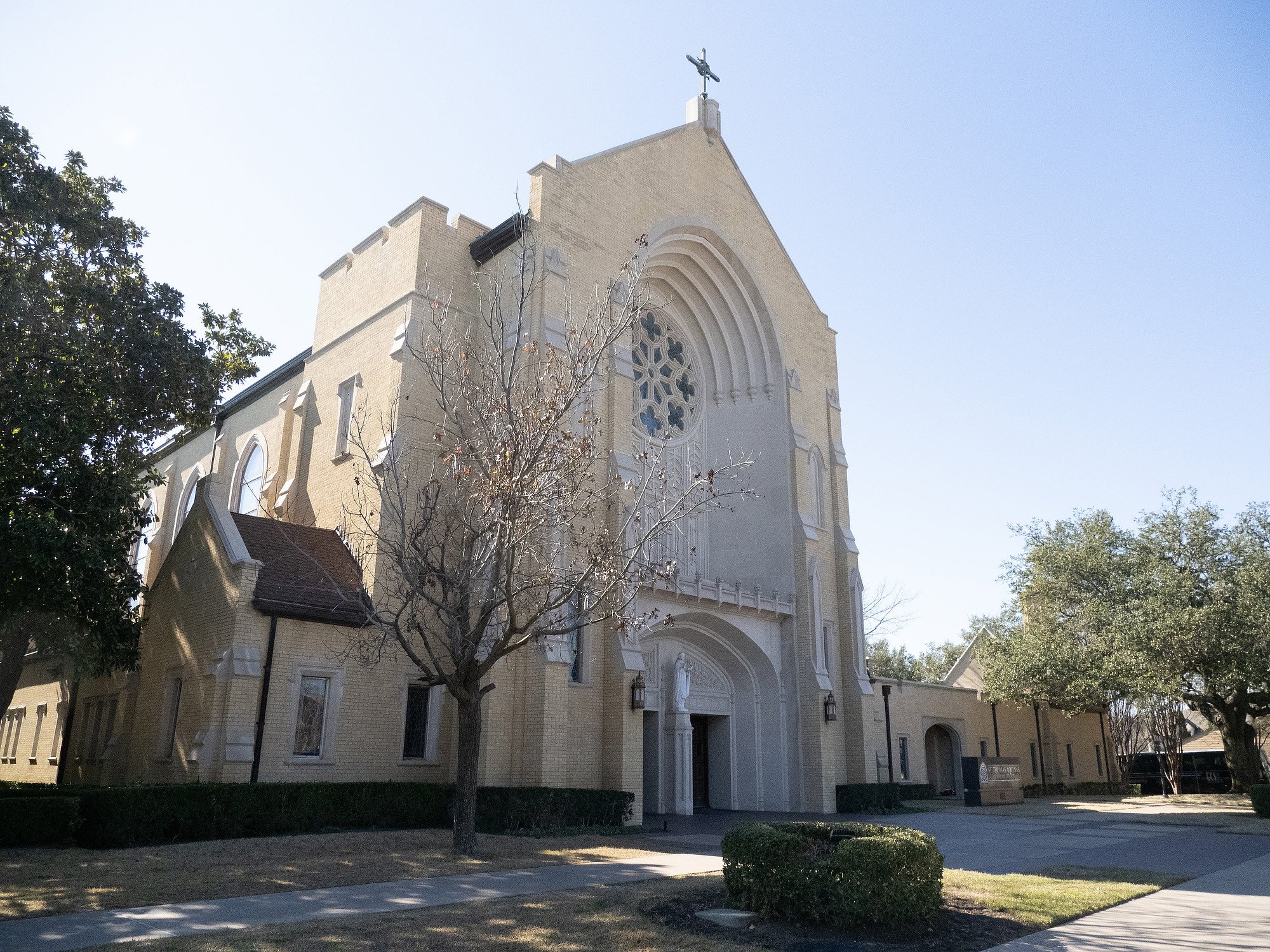Exterior view of a church with a cross on the roof, tall arched windows, trees, well-kept lawn, and a walkway leading to the entrance.