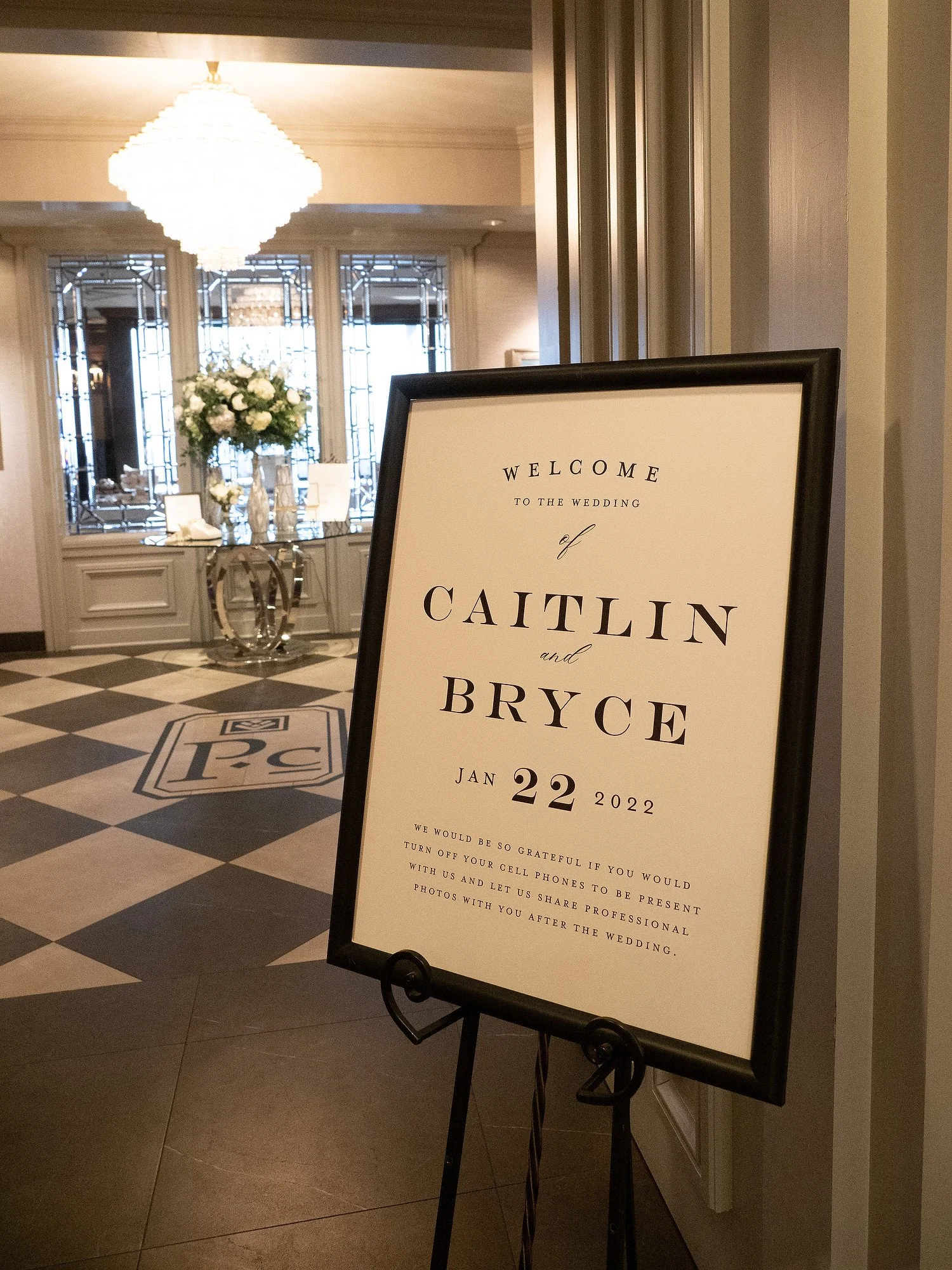 Wedding welcome sign with the names Caitlin and Bryce, February 22, 2022, in an elegant venue with floral arrangements and a chandelier in the background.