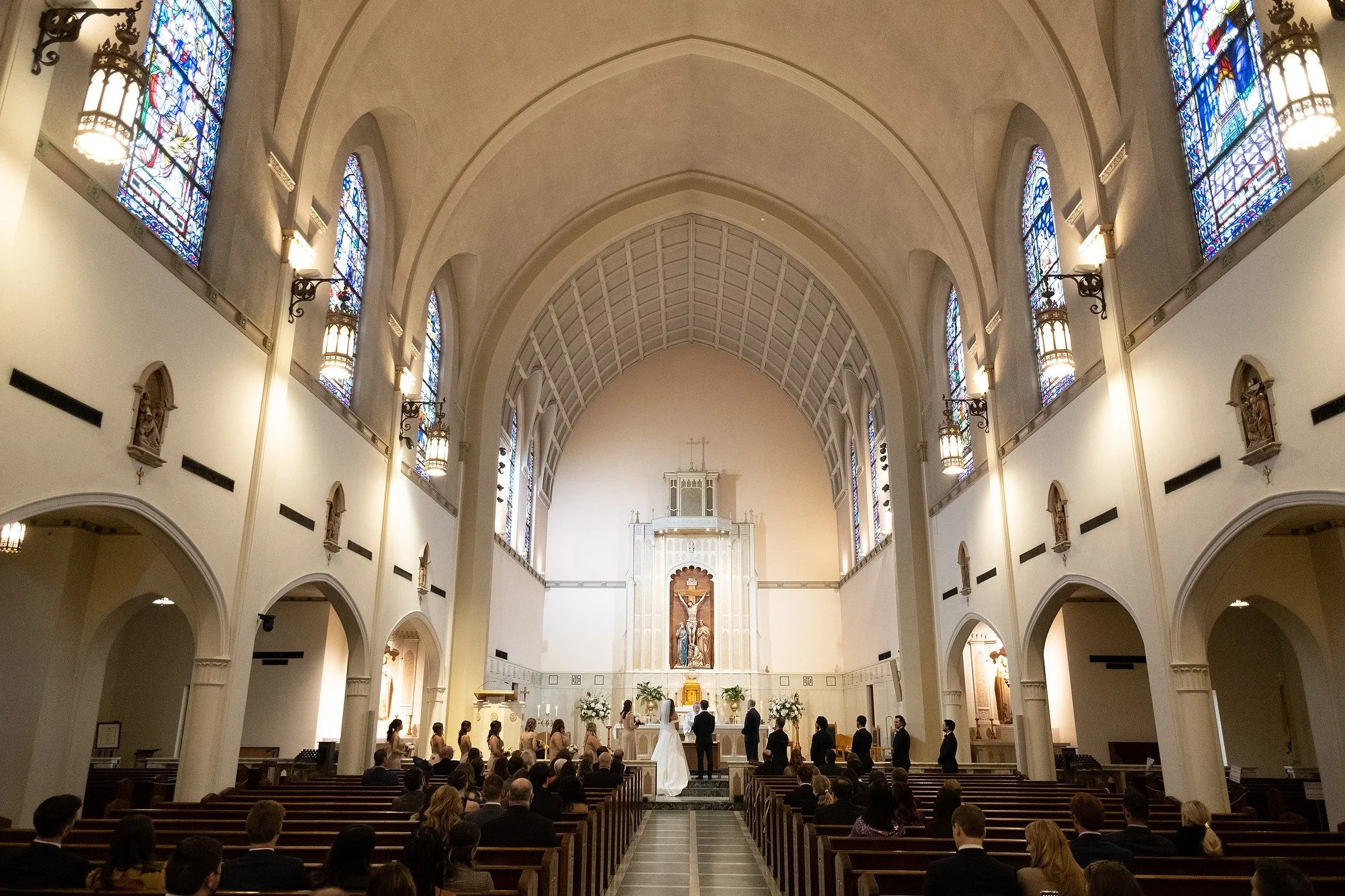 Inside a church during a wedding ceremony with the altar, religious statues, stained glass windows, and seated guests.