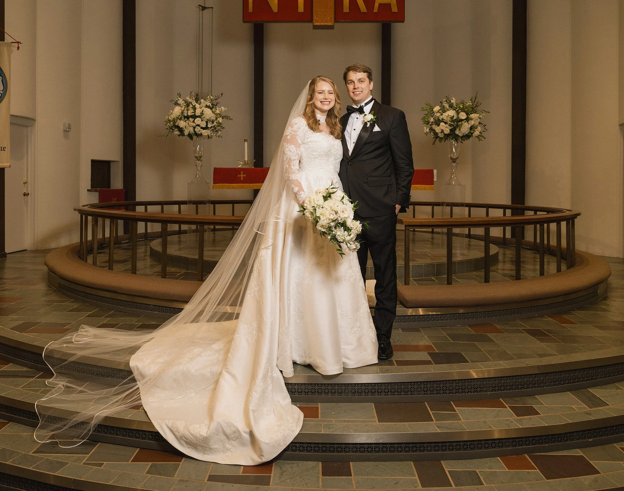 Bride and groom standing together on a church altar during their wedding ceremony, smiling at the camera, with flowers and religious symbols in the background.