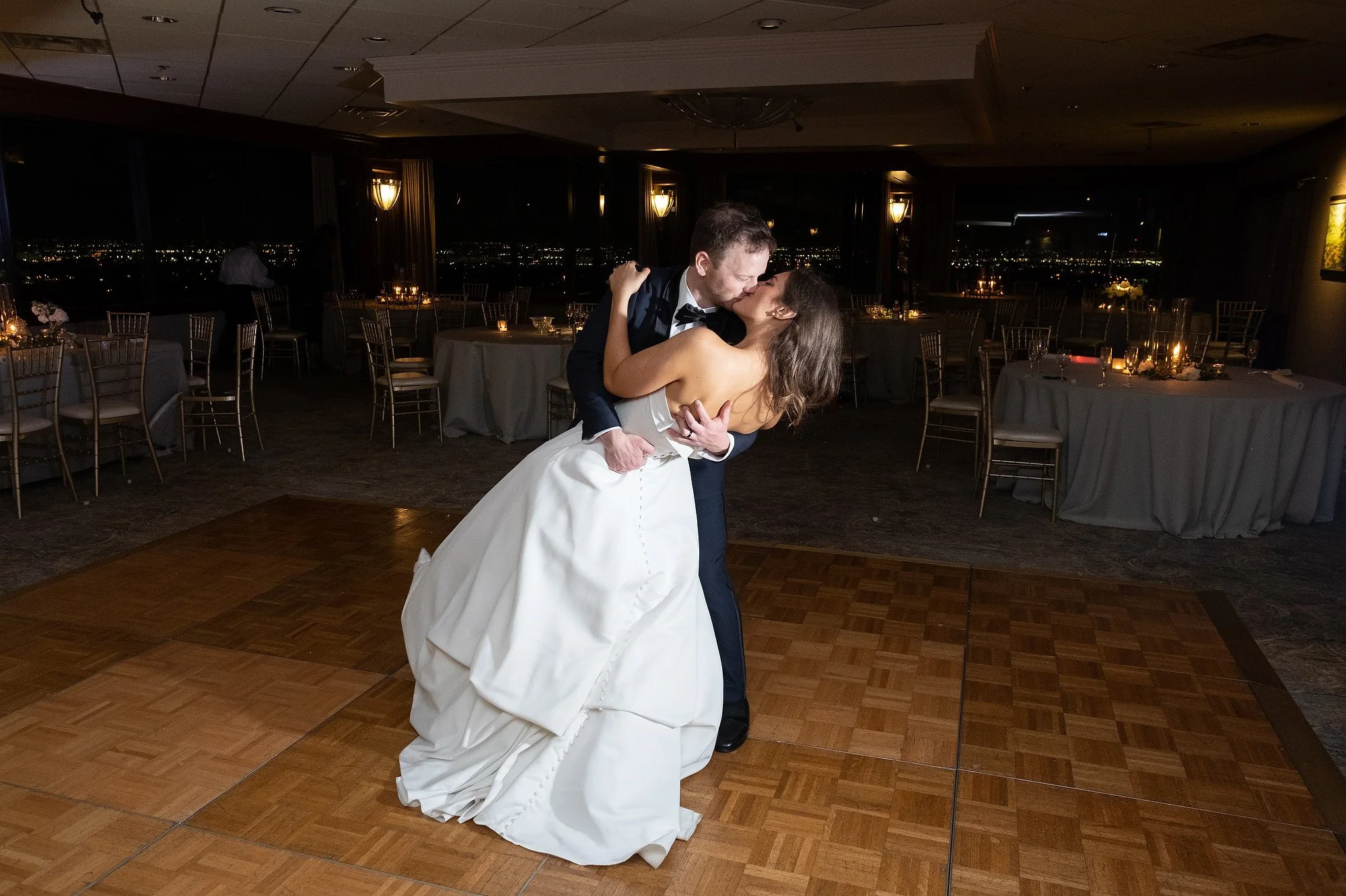 A couple in wedding attire sharing a dance on the dance floor at night, with a city view through the windows and decorated tables around them.