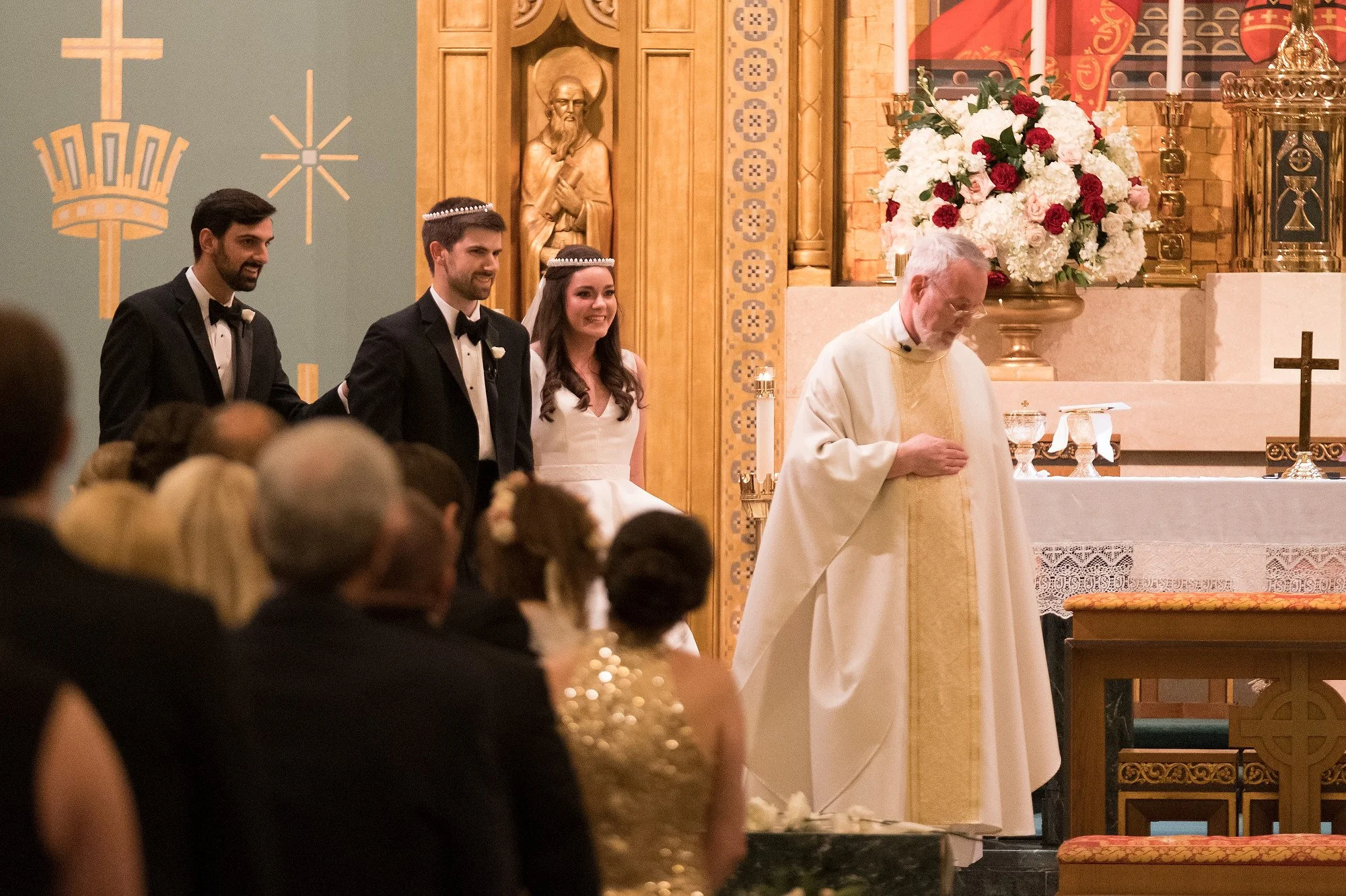 A wedding ceremony taking place in a church with three bridesmaids and a priest. The groom and bride are smiling, dressed in tuxedos and a white wedding dress respectively. The background features religious symbols, a statue, and a large floral arran