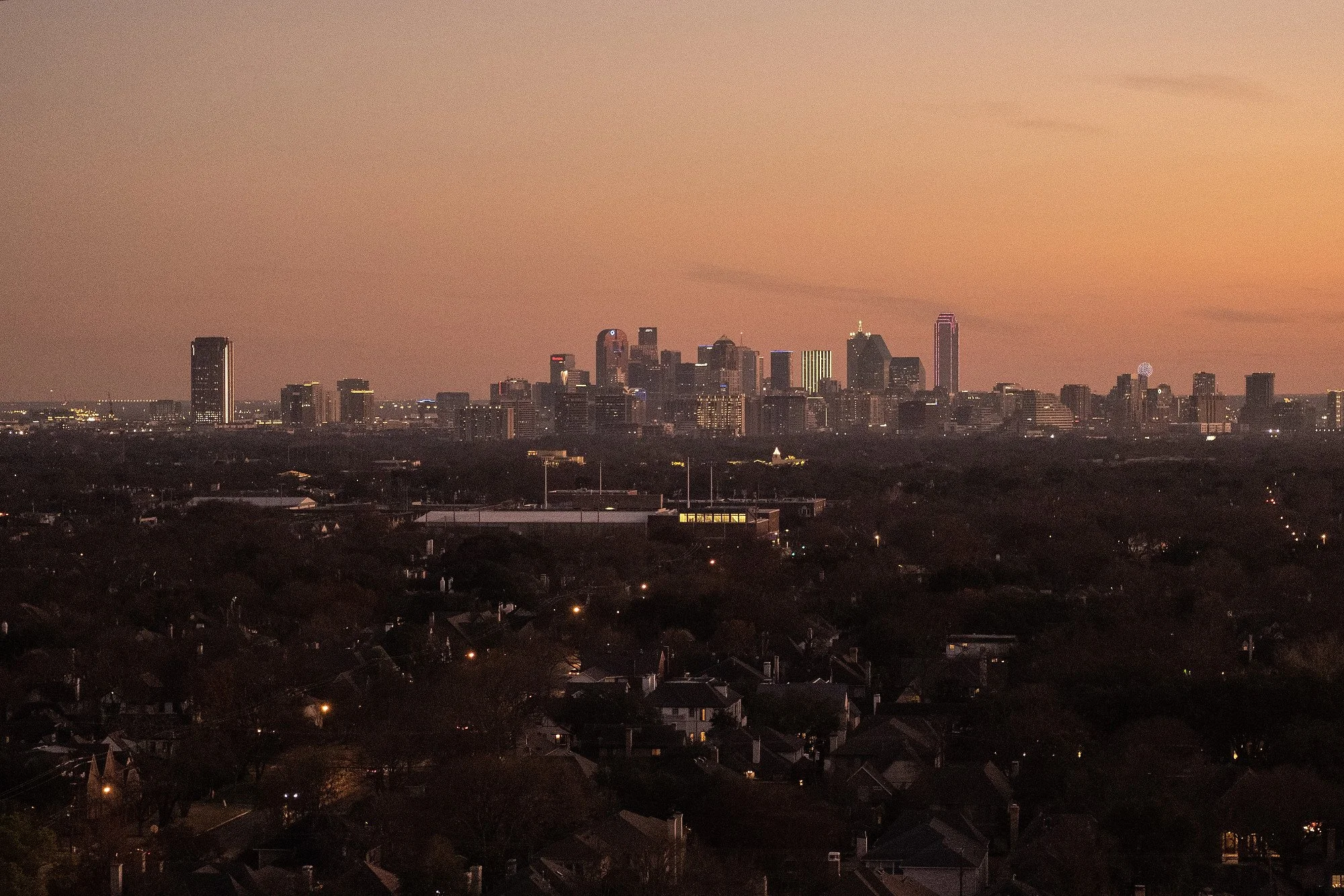 City skyline at dusk with tall buildings and a gradient orange and purple sky, viewed over residential neighborhoods.