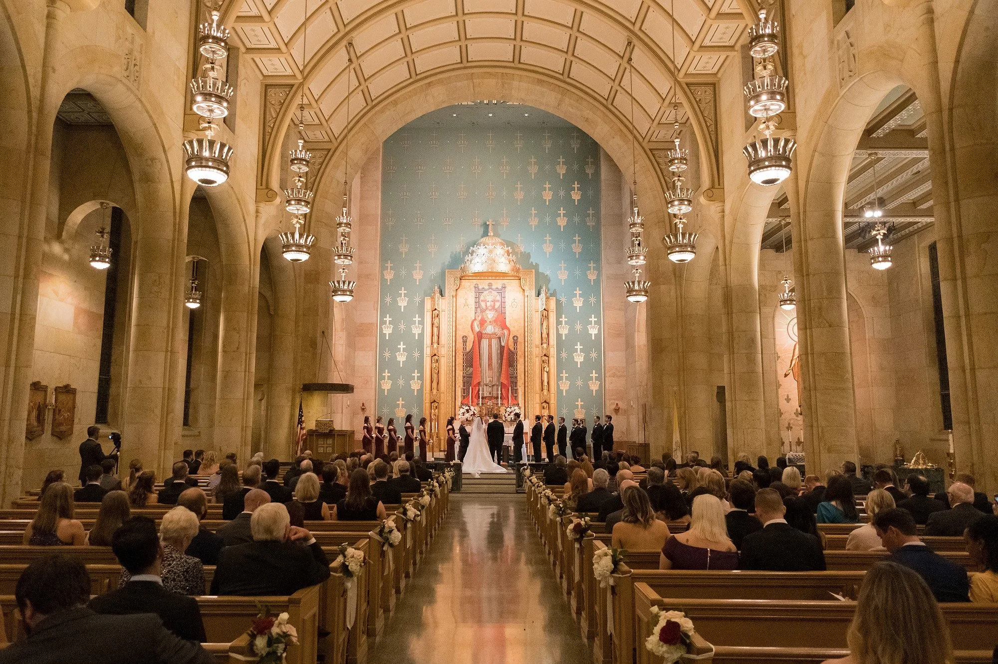 A wedding ceremony taking place inside a grand church with high arched ceilings, ornate lighting, and a large altar featuring religious artwork. The bride and groom stand at the altar surrounded by clergy, with guests seated in pews watching the cere