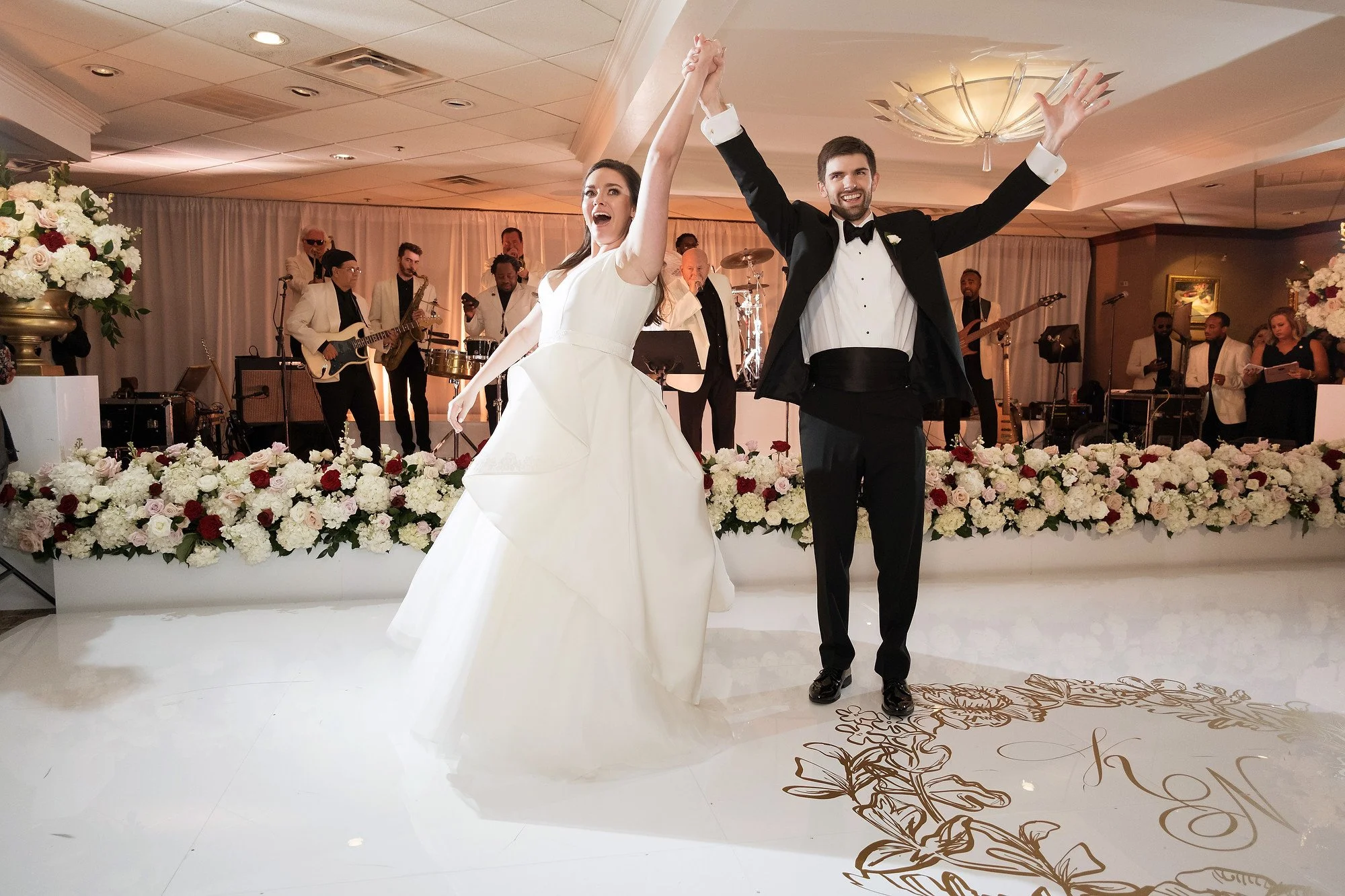 A newlywed couple dancing together at their wedding reception, with a band playing live music in the background and floral decorations on the stage.