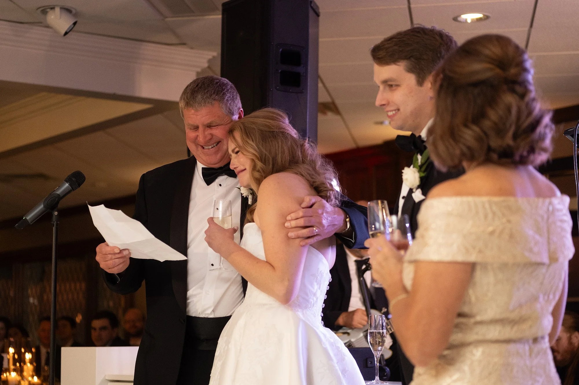 A bride and groom sharing a moment of laughter and hugging during their wedding toast with two other guests, while a man reads a speech at the microphone.