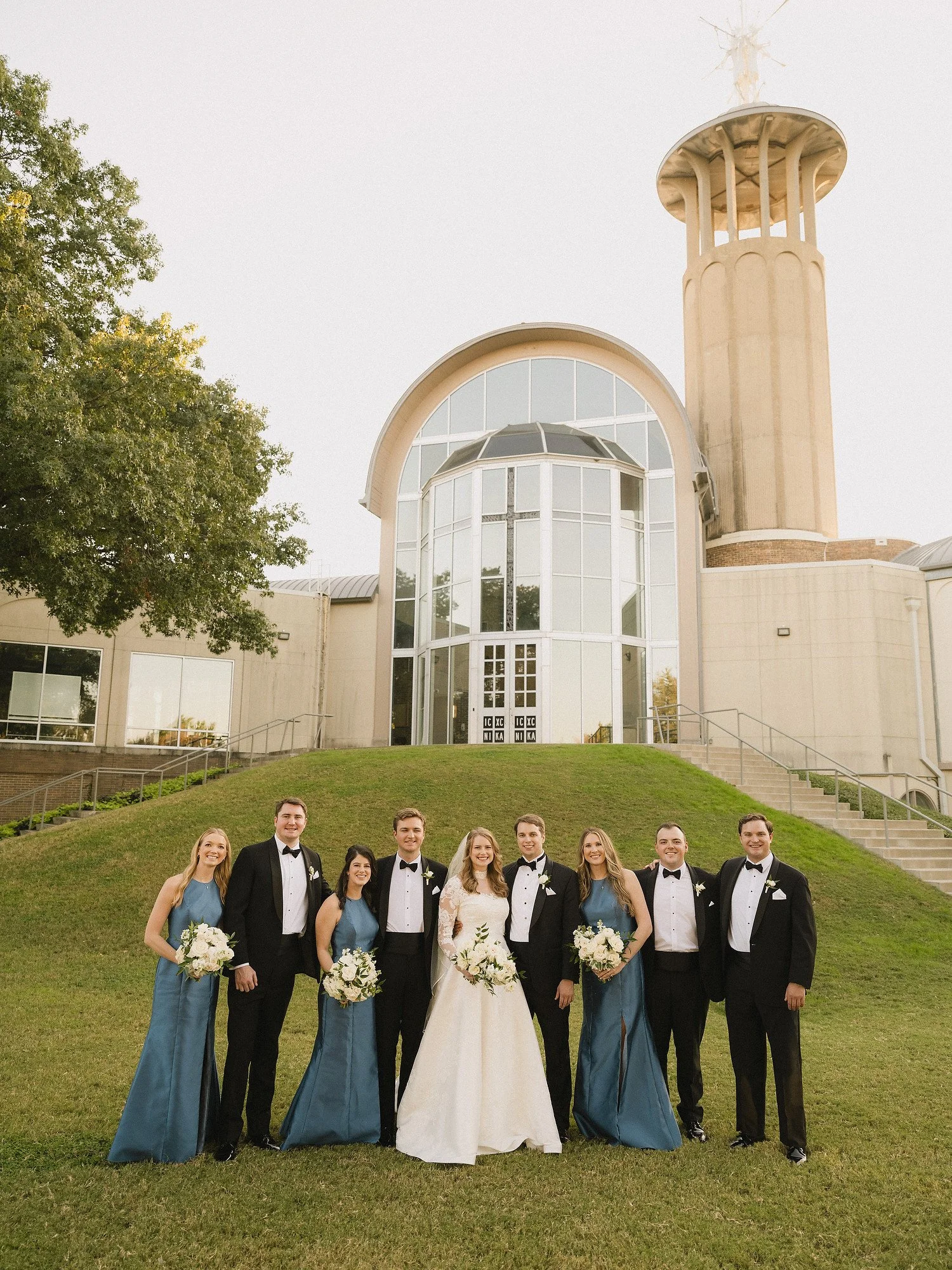 Group of wedding party members, including bride, bridesmaids, and groomsmen, standing on a grassy hill in front of a church with modern architecture and a tall steeple. The bride is in a white gown, the bridesmaids wear blue dresses, and the groomsme