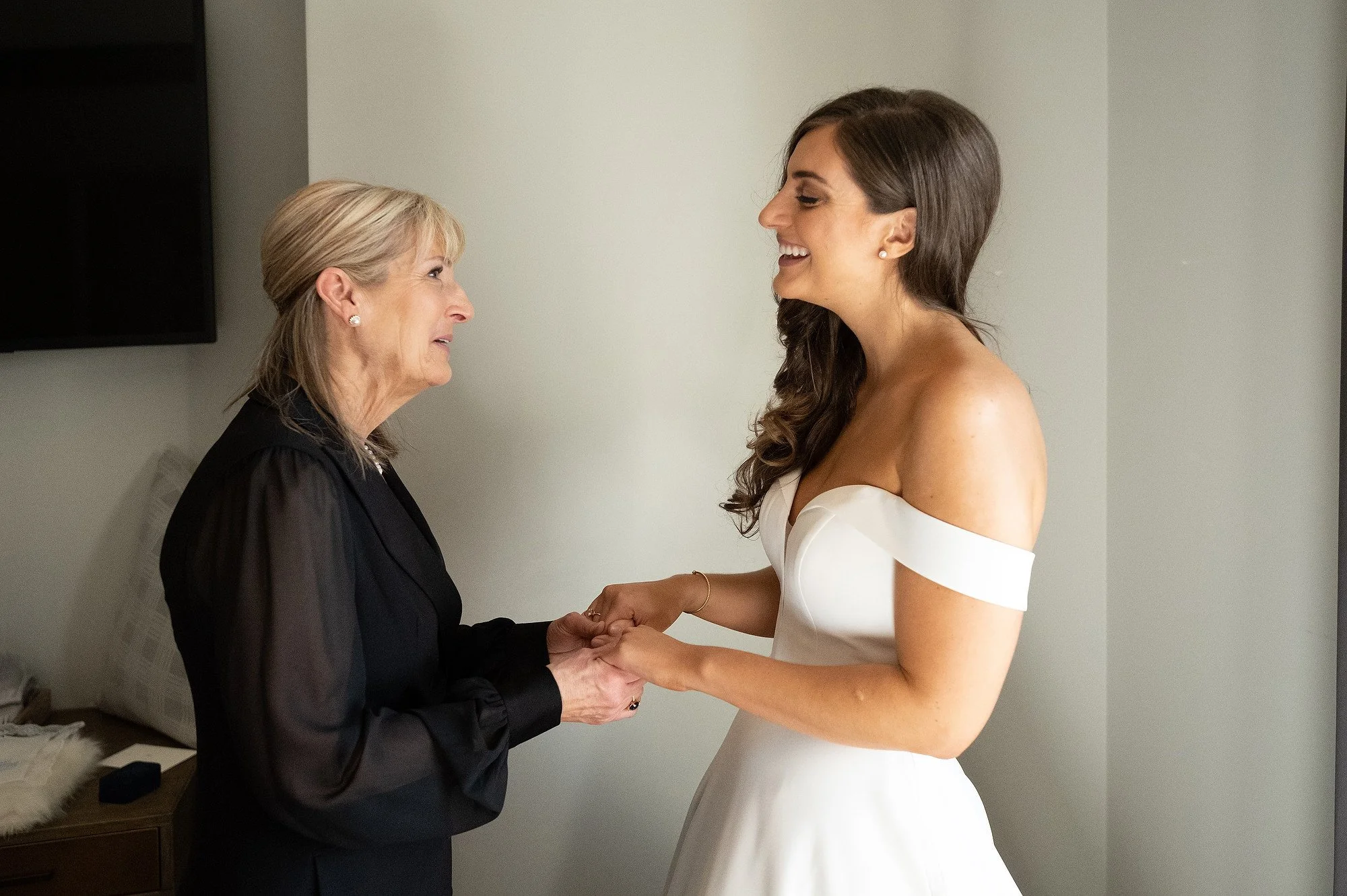 A young woman in a white off-shoulder dress holding hands with an older woman in black, both smiling and looking at each other, in a light-colored room.
