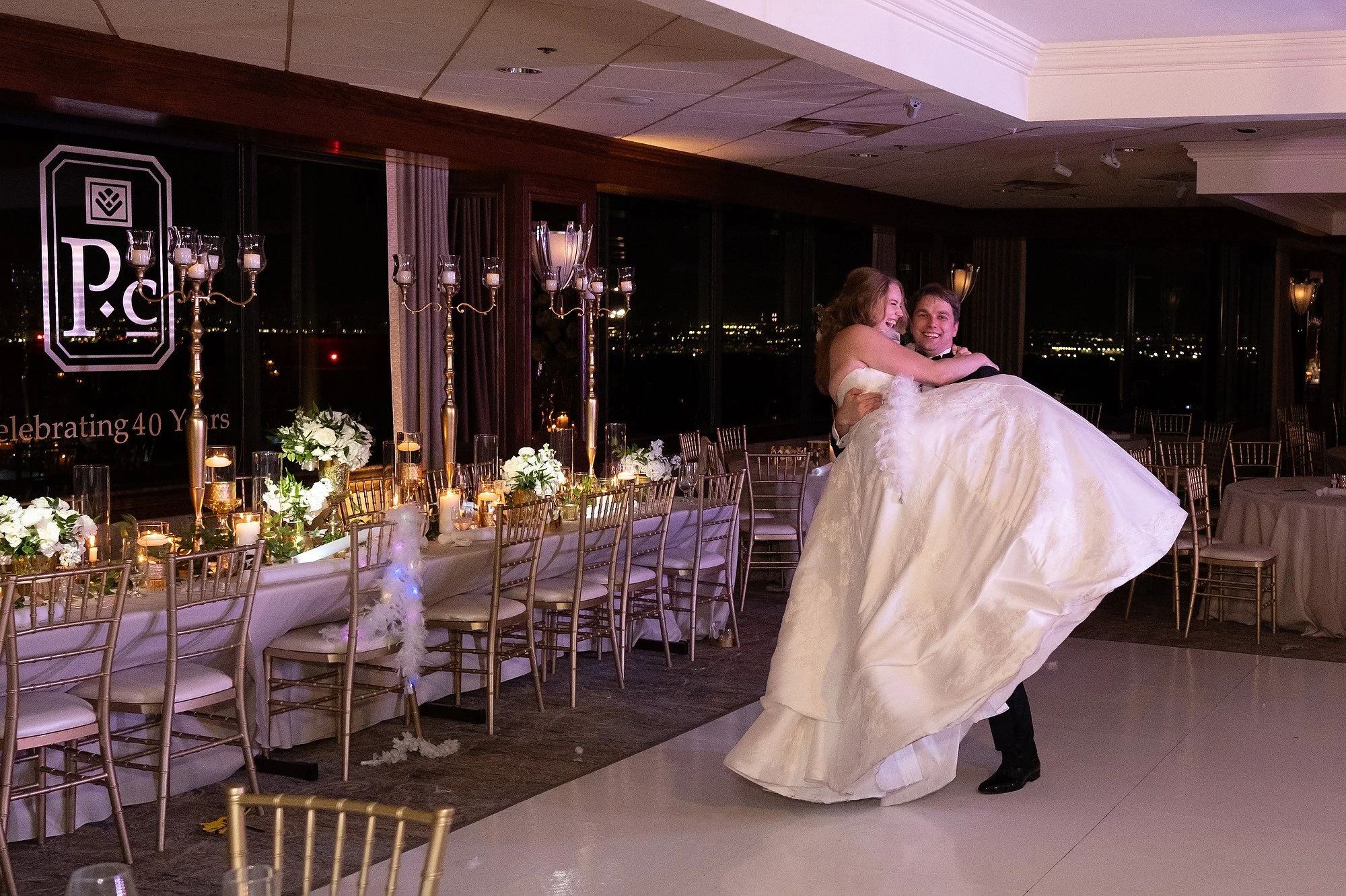 A bride being lifted by a groom on a dance floor at a wedding reception, with elegant table settings featuring white flowers, candles, and tall candelabras in the background.