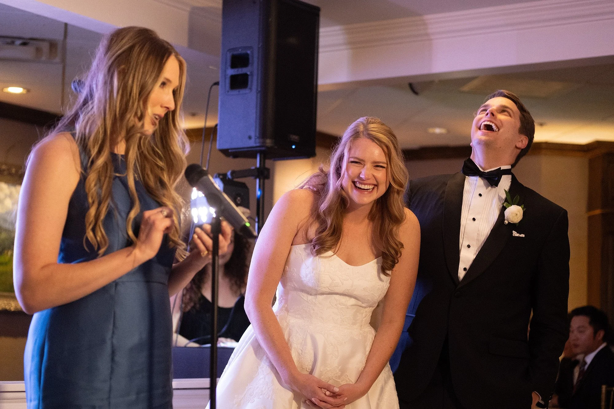 A bride and groom stand together, laughing happily during their wedding reception, while a woman in a blue dress reads vows or a toast.