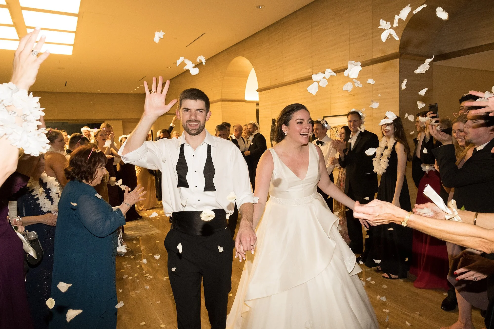 A newlywed couple walks hand-in-hand through a crowd of wedding guests throwing flower petals in the air inside a warmly lit banquet hall.
