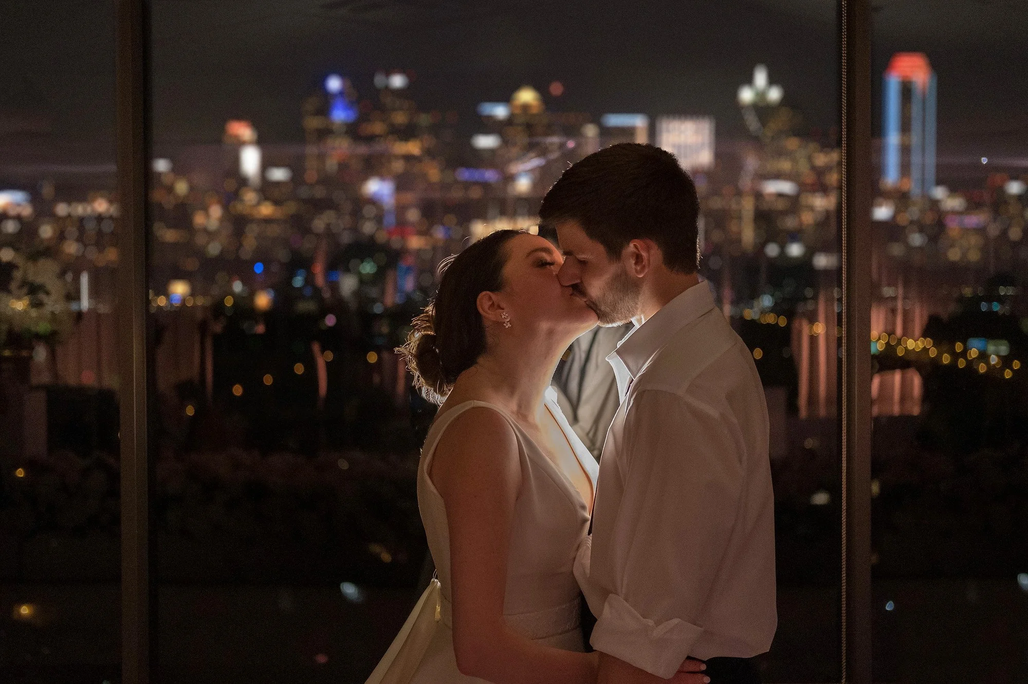 A couple sharing a kiss at night, standing close by a window with a city skyline in the background.