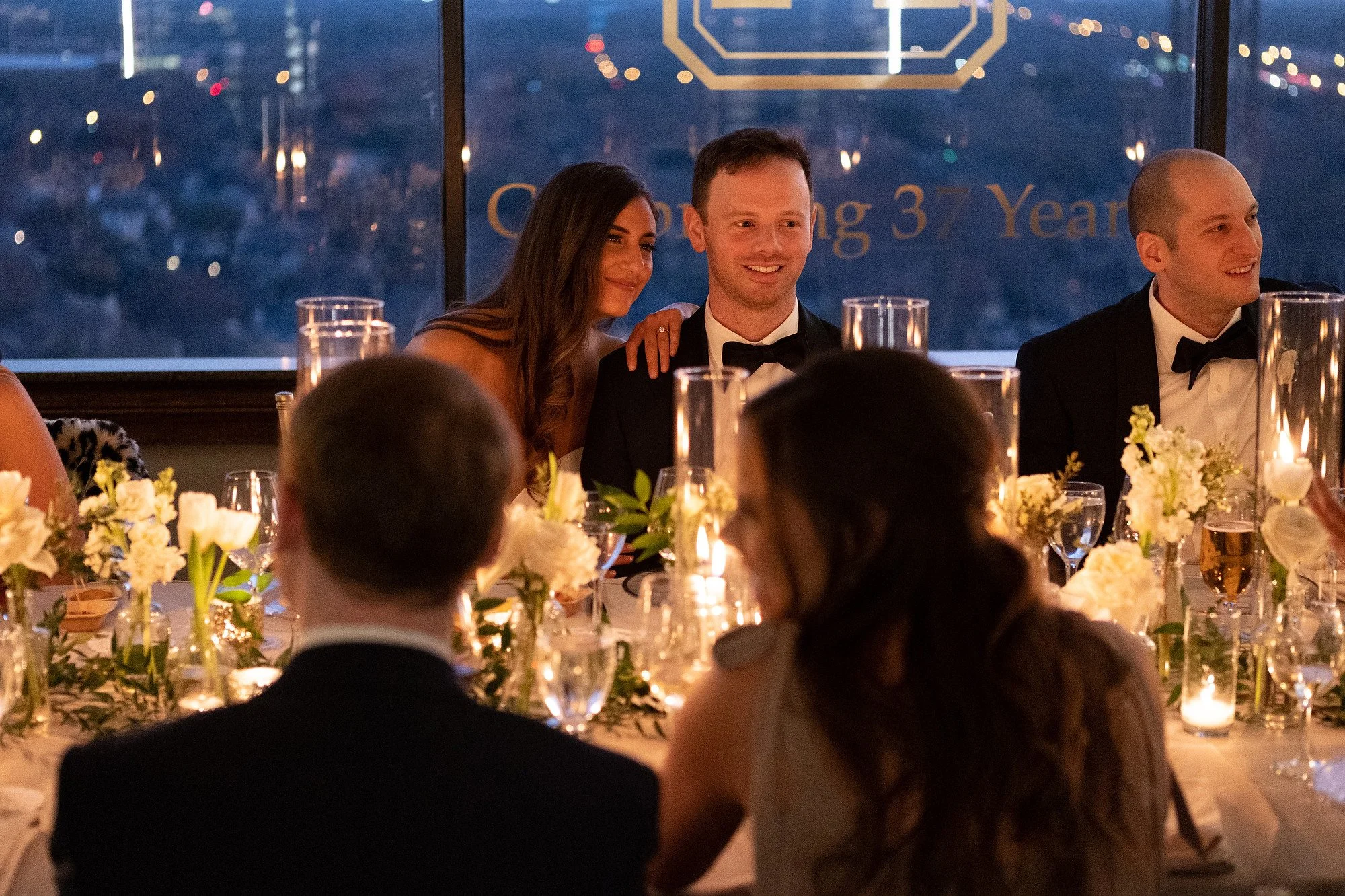 A wedding reception with a bride and groom sitting at a decorated table, smiling, with other guests in the foreground. The background features a cityscape view through large windows.