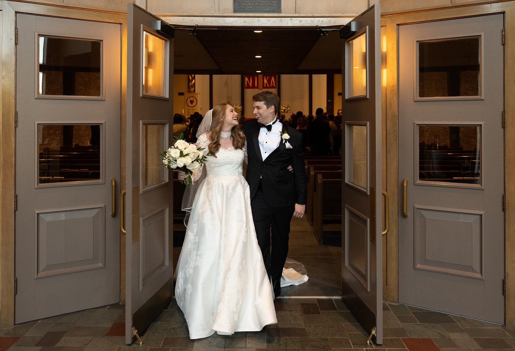 A bride and groom smiling and looking at each other as they walk through church doors after their wedding, with guests visible inside.