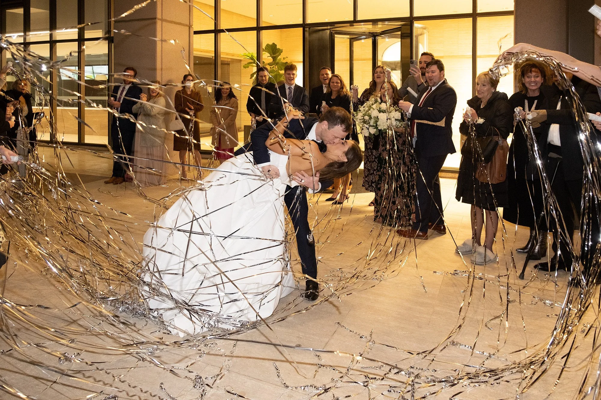 A bride and groom kissing amid shower streamers at their wedding reception, surrounded by seated and standing guests taking photos and celebrating inside a modern venue.