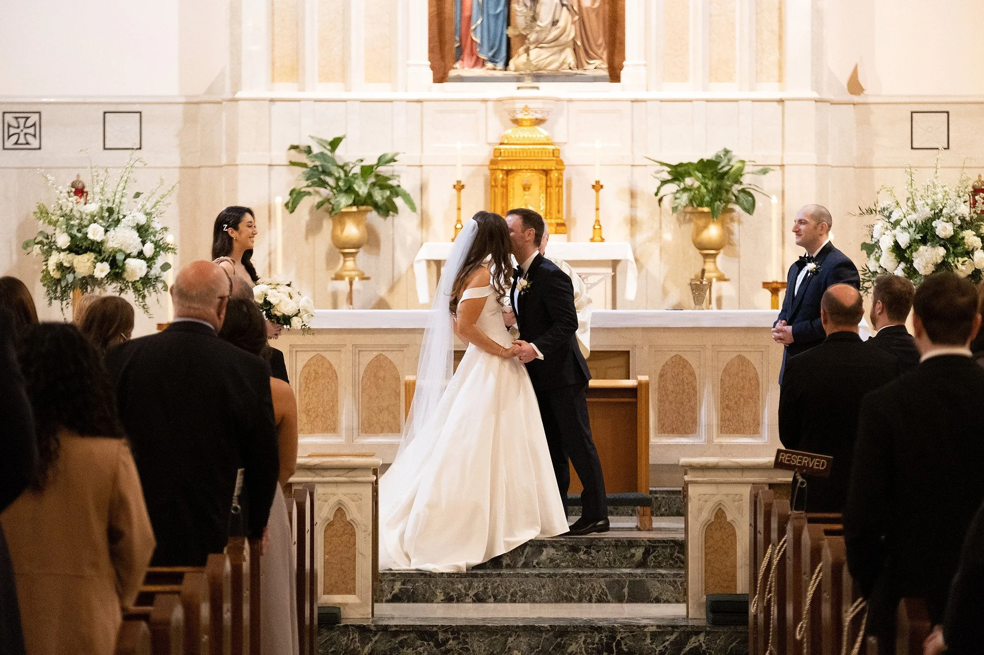 Wedding ceremony in a church with a bride and groom kissing at the altar, surrounded by bridesmaids, groomsmen, and guests seated in pews, during the marriage vows.